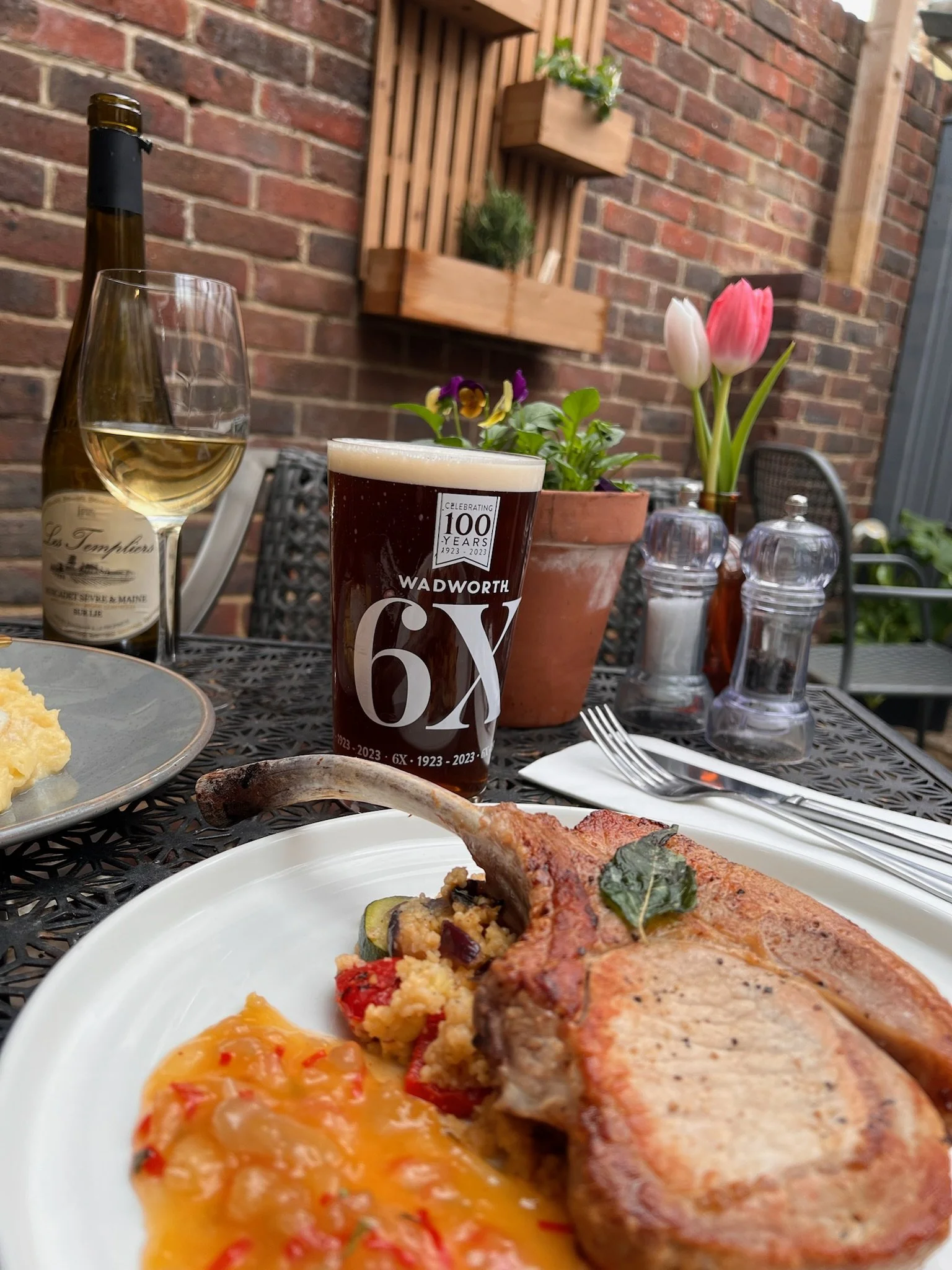 Outdoor dining table with a plate of pork chop, mashed potatoes, and vegetables, a glass of white wine, a pint of dark beer, a bottle of white wine, pink and white tulips, salt and pepper shakers, and lettuce in a flowerpot, set against a brick wall.