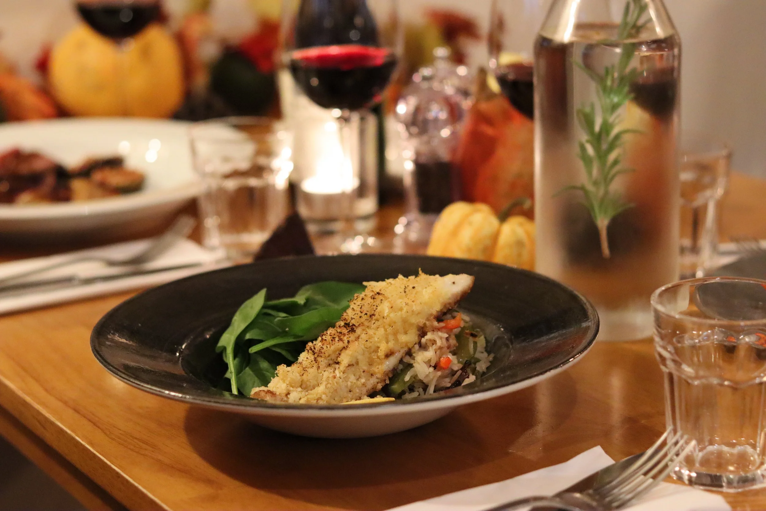 A plated serving of baked white fish with a breadcrumb topping, served over a bed of greens and vegetables, on a black and white plate on a wooden table with wine glasses and a water pitcher in the background.