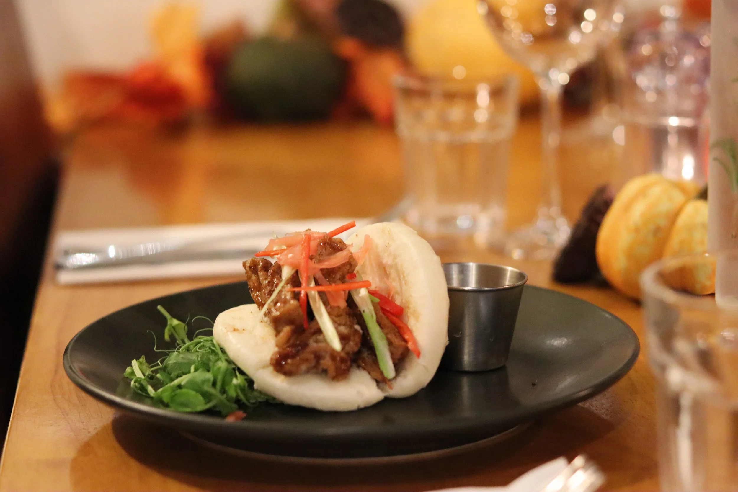 A bao bun filled with tender beef and topped with julienned vegetables, served on a black plate with green garnish and a small metal cup of sauce, on a wooden table with glasses and bread in the background.