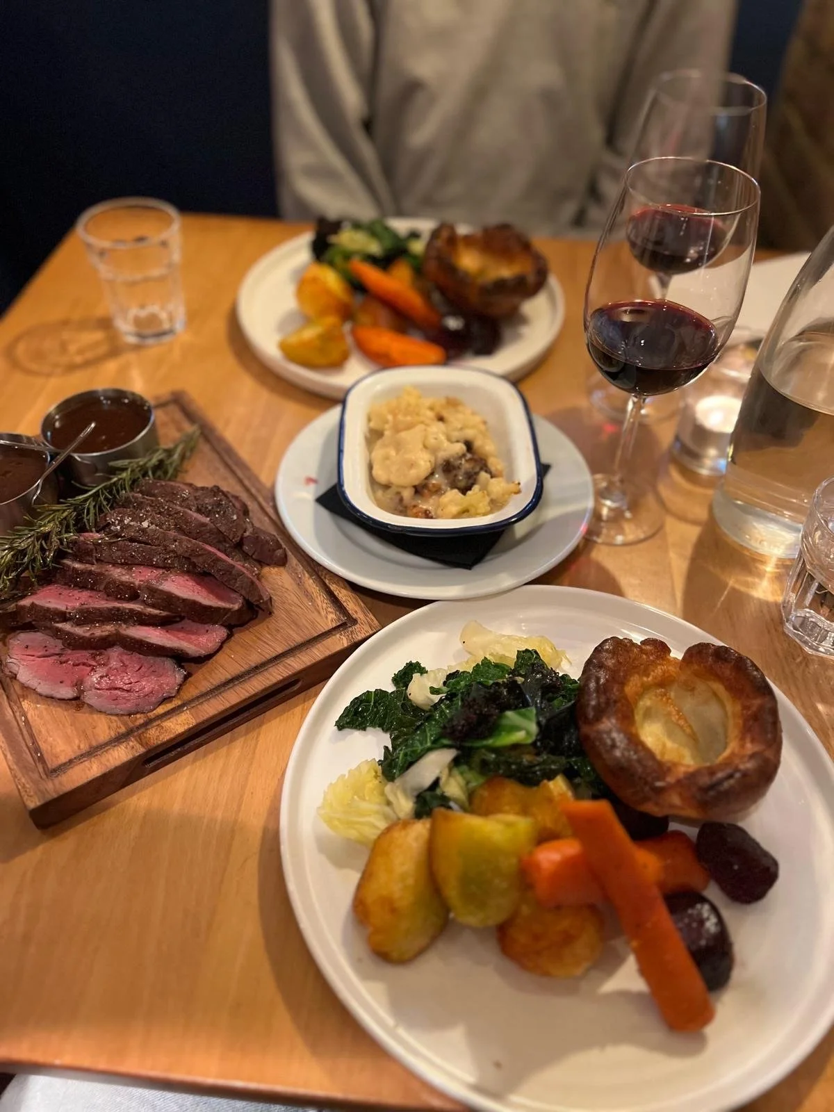 A dinner table with a plate of cooked beef, roasted vegetables, a small dish of mashed potatoes, a biscuit, glasses of red wine, and water, along with other dishes of vegetables and roast on the table.