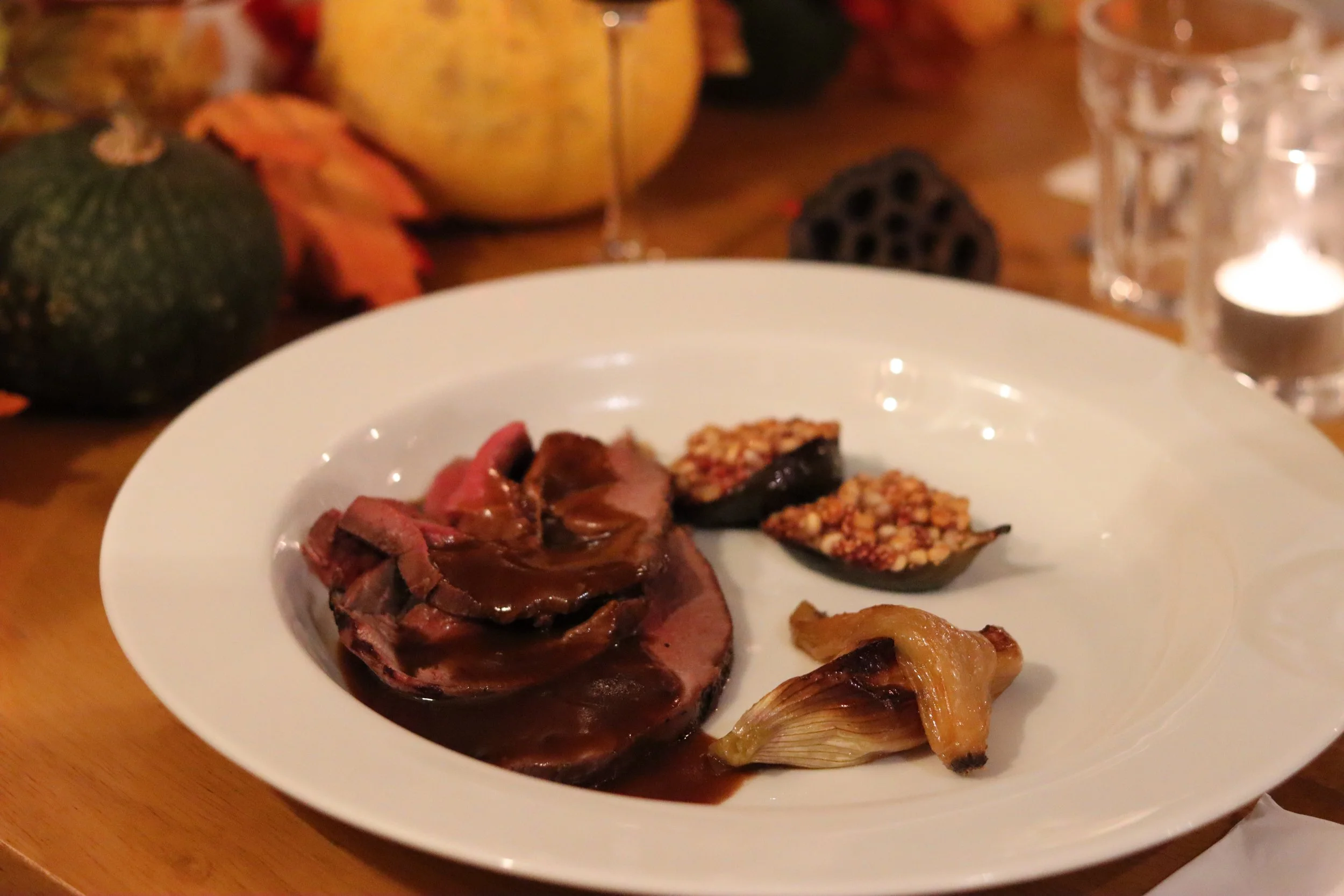 A white plate with roast beef, stuffed figs, roasted garlic, and sweet potato casserole, set on a wooden table with pumpkins, leaves, a glass of wine, and candles in the background.