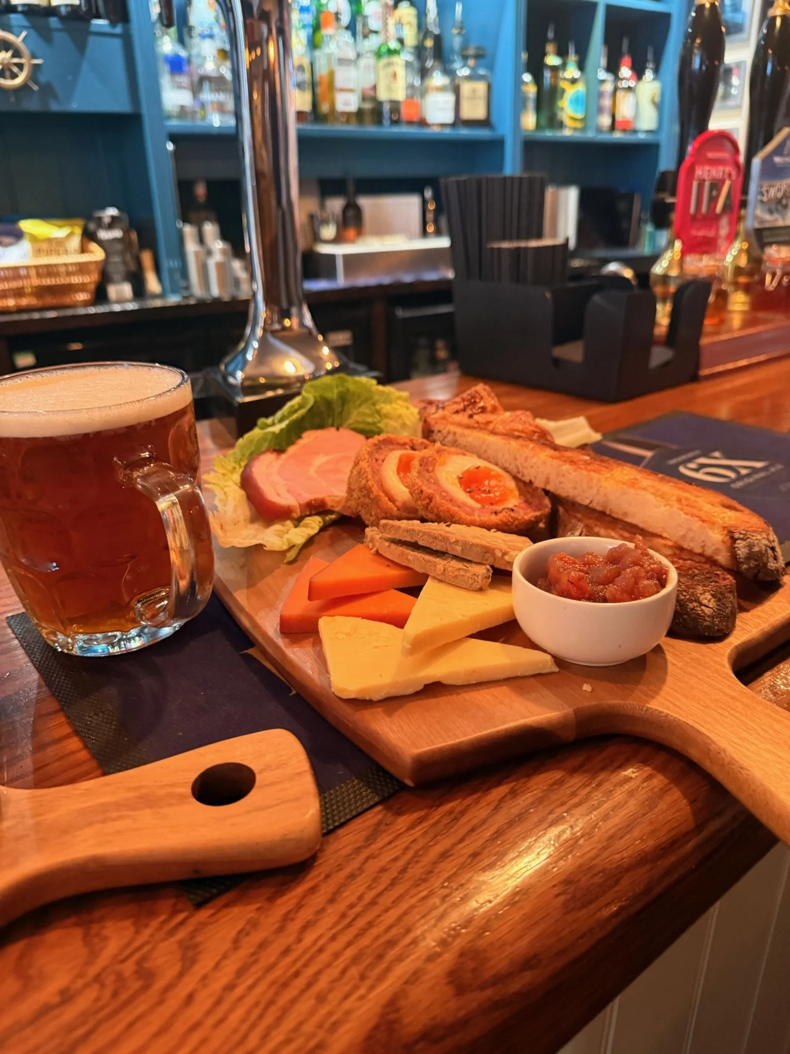 A wooden serving board with various cheeses, sliced meats, bread, and a small bowl of chutney, accompanied by a mug of beer, on a bar counter with liquor bottles in the background.