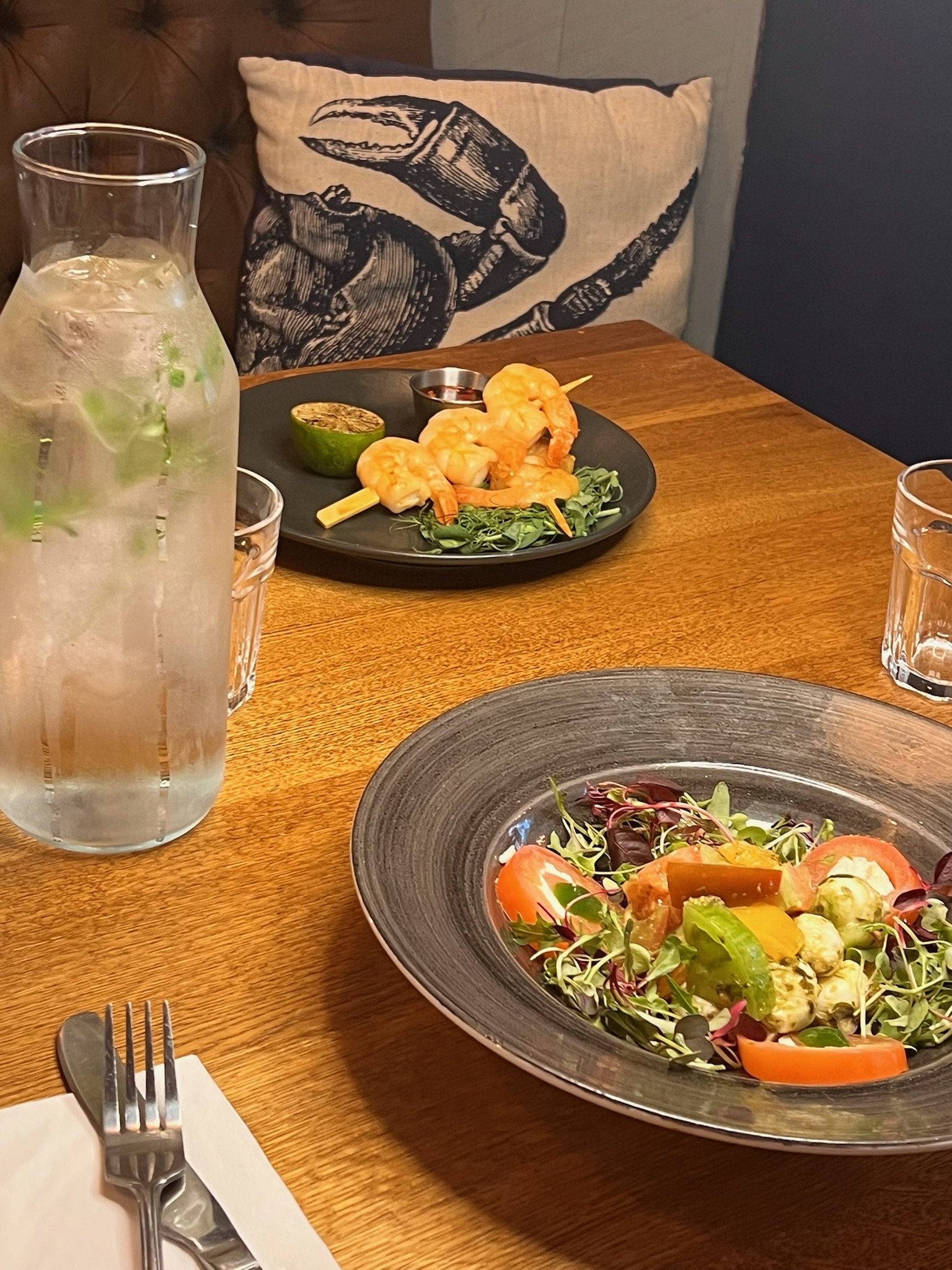 A table with a large glass pitcher of water, a salad with tomatoes and greens, and a black plate with cooked shrimp, lime, and dipping sauce. In the background, a decorative pillow with a crab illustration is visible.
