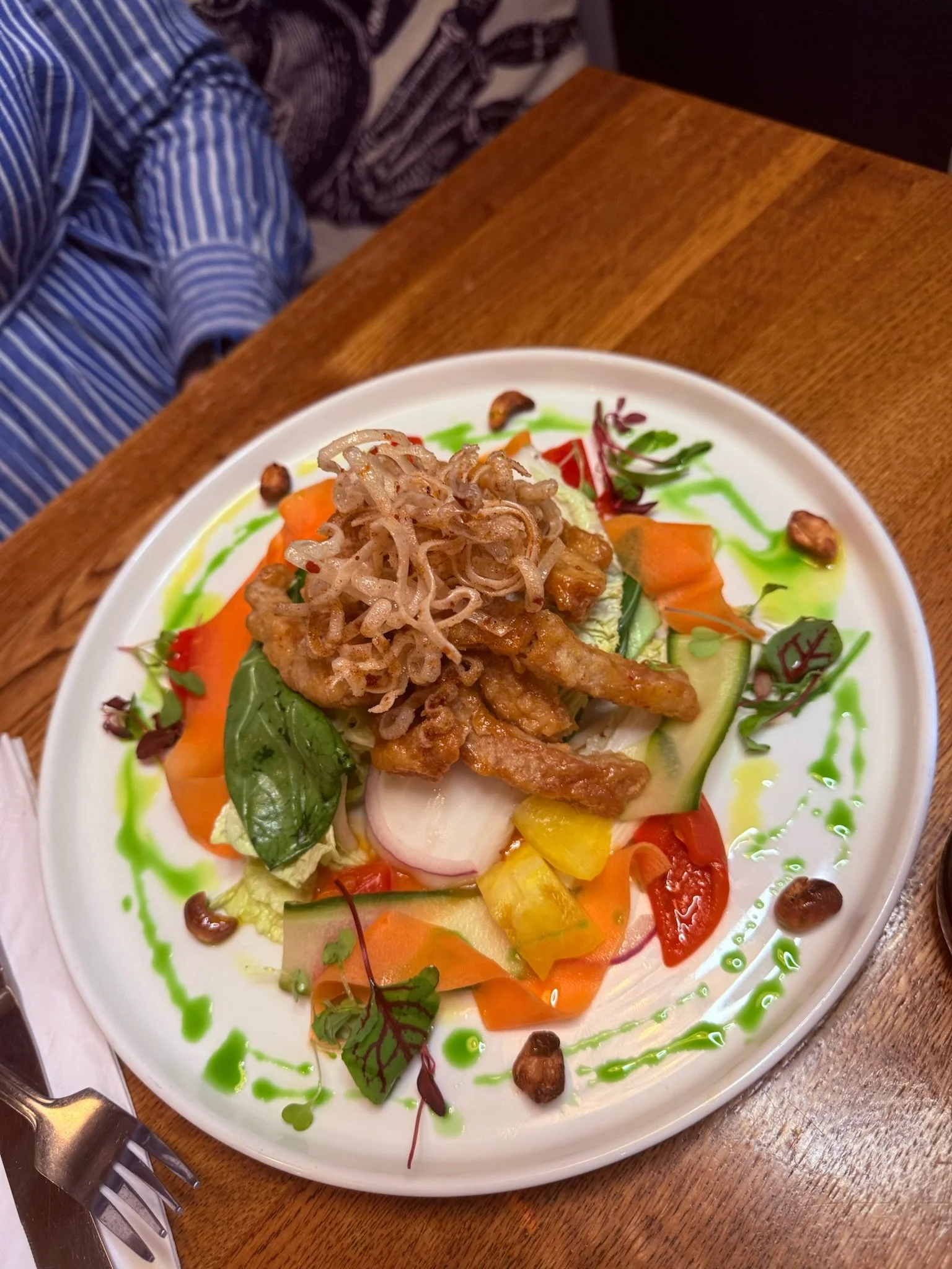 A colorful salad on a white plate with mixed greens, sliced cucumbers, tomatoes, radishes, yellow peppers, and roasted nuts. Topped with crispy fried noodles and drizzled with green dressing, served on a wooden table.