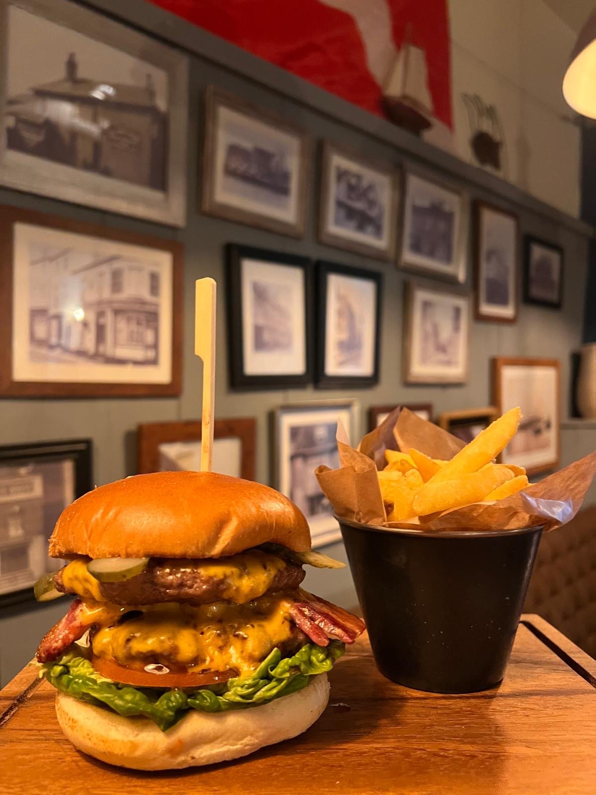 A large cheeseburger with lettuce, tomato, pickles, bacon, and two beef patties on a bun, served with a side of French fries in a black cup, on a wooden table in a restaurant with framed pictures on the wall.