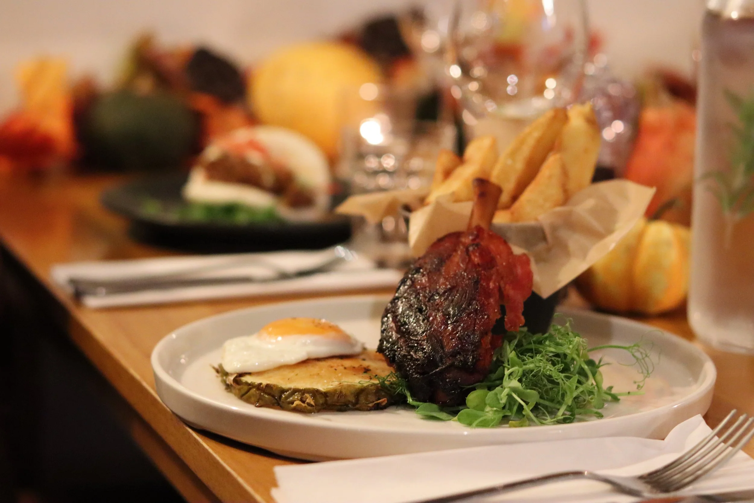 Plate with baked potato topped with a fried egg, grilled meat, and green garnish, with a side of French fries in paper wrapper.