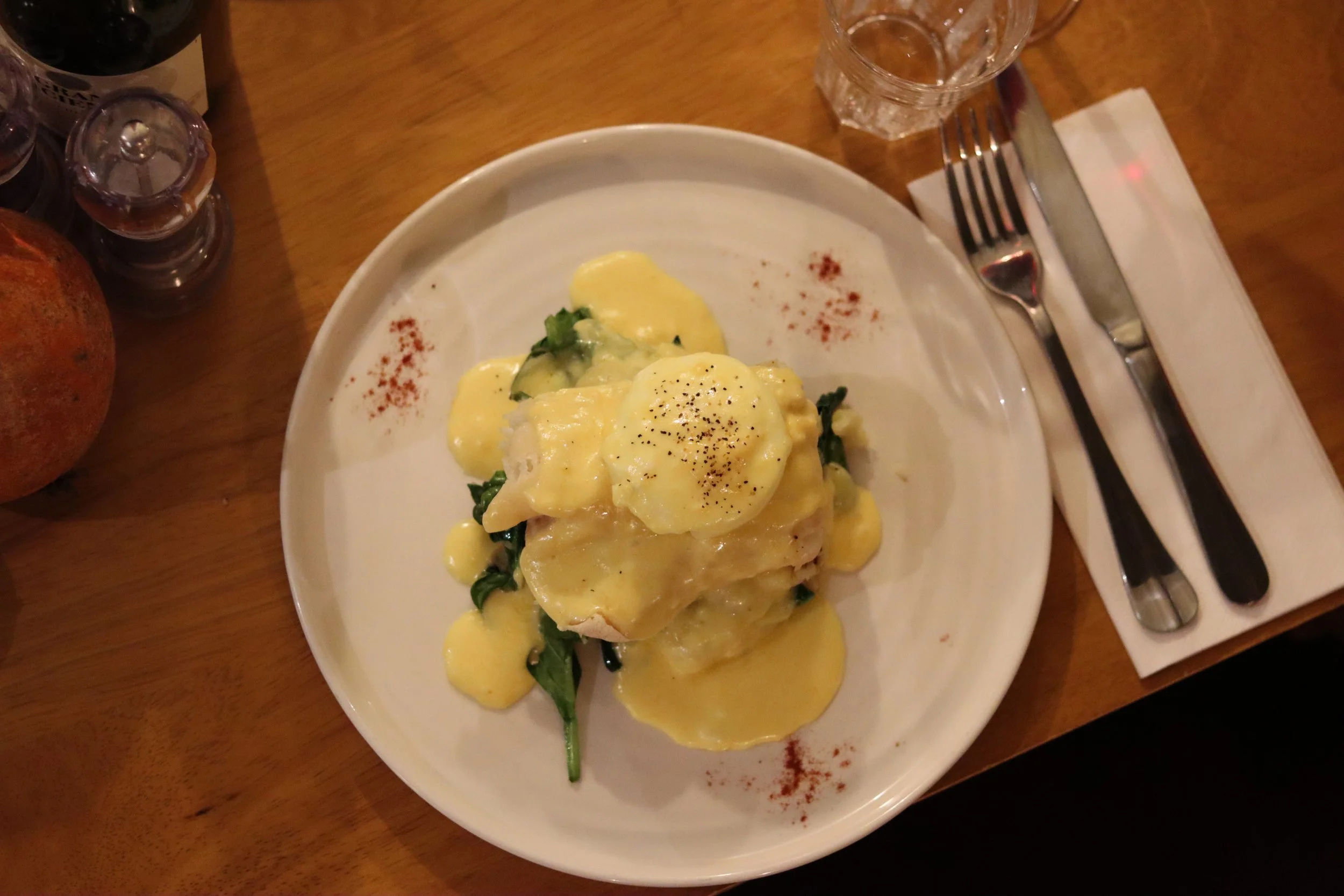Plate of eggs Benedict topped with hollandaise sauce, served with spinach and sprinkled with black pepper, on a white dish. Side of red pepper flakes on the plate. Beside a knife, fork, and napkin on the right. Glass of water above the plate. Wooden 
