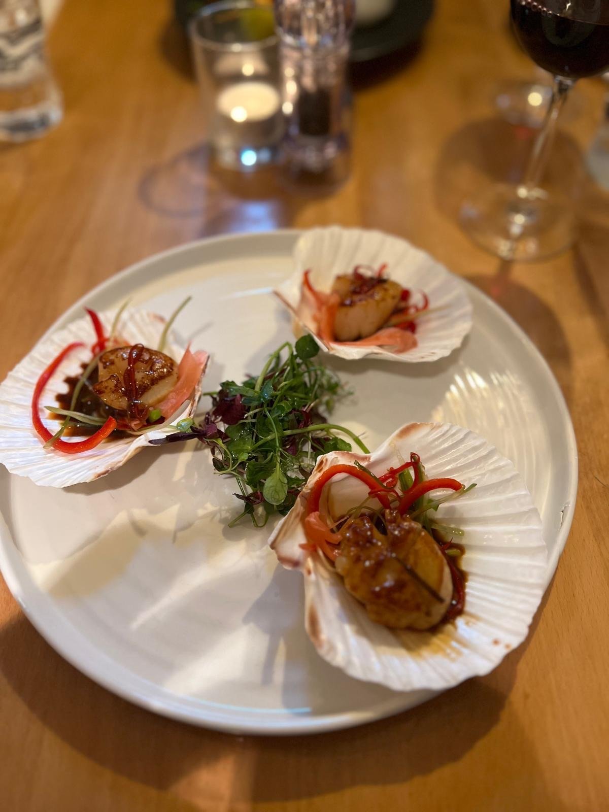 Three stuffed scallop shells topped with sauce and garnished with microgreens on a white plate, with glasses of red wine and water in the background.