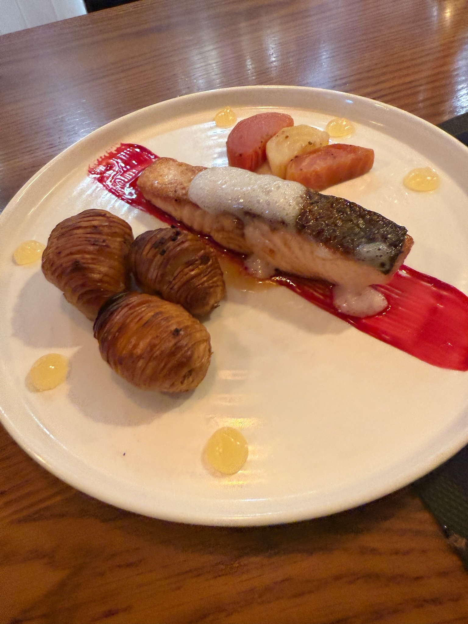 A white plate with a cooked fish fillet topped with foam and a red sauce smear, three roasted snail shells, and slices of sausage, onions, and potatoes with yellow sauce dots around.