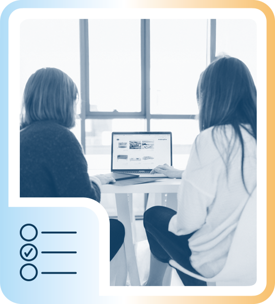 Two women sitting at a table looking at a laptop screen in a modern office with large windows.