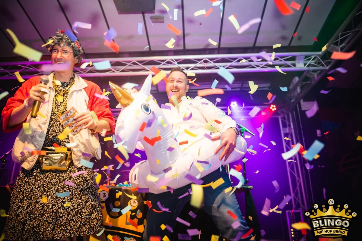 Two men on stage celebrating with colorful confetti, one holding a microphone and the other sitting on an inflatable unicorn float, smiling amidst party lights.