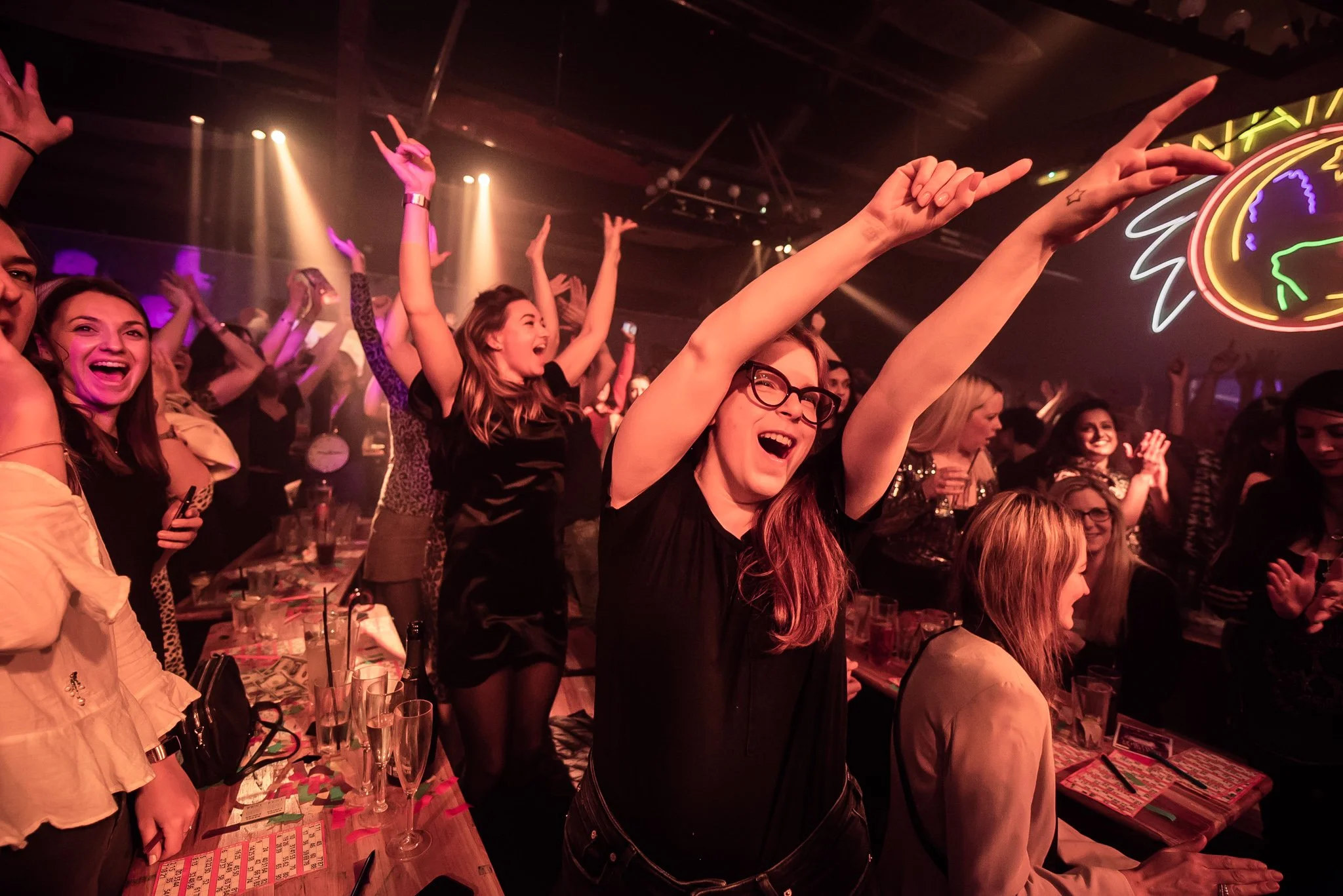 A lively crowd of young women at a hip hop bingo night in Portsmouth, dancing and smiling with neon lights in the background.