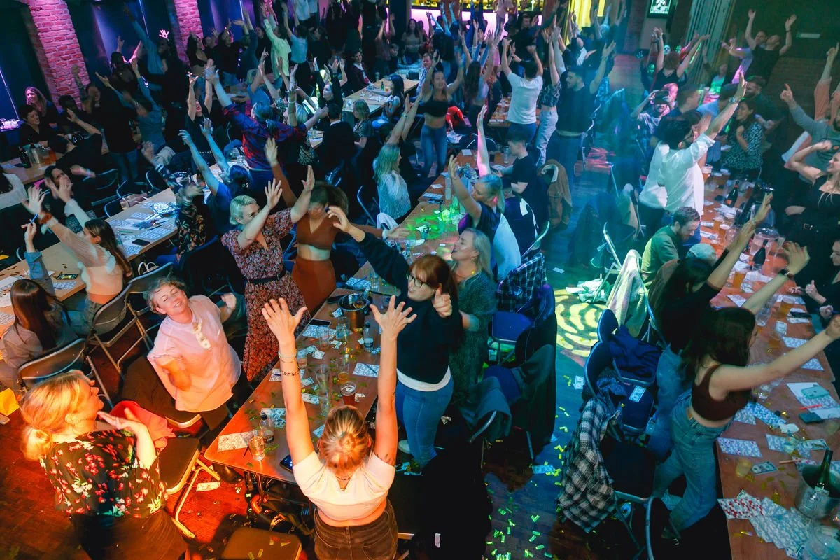 A large group of people gathered, dancing, and raising their hands after a blingo bingo show. The tables are filled with drinks, and the atmosphere is lively with colourful lighting.