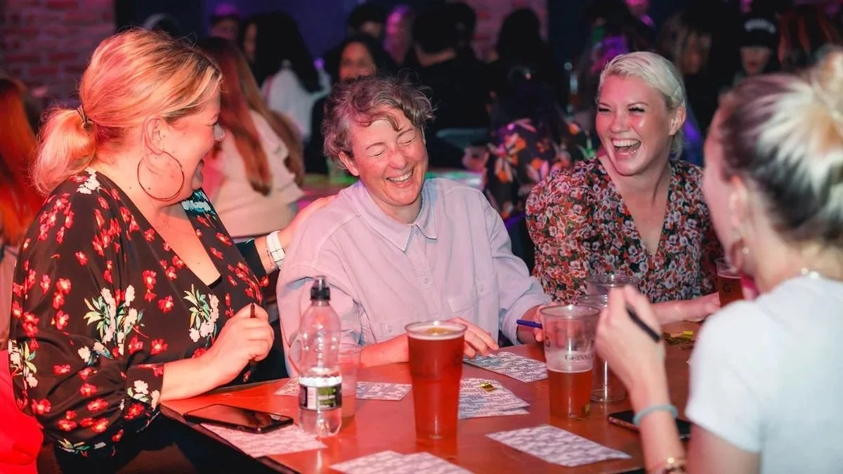 A group of people sitting at a table, laughing and enjoying drinks together at Hip Hop Bingo with colourful lighting.