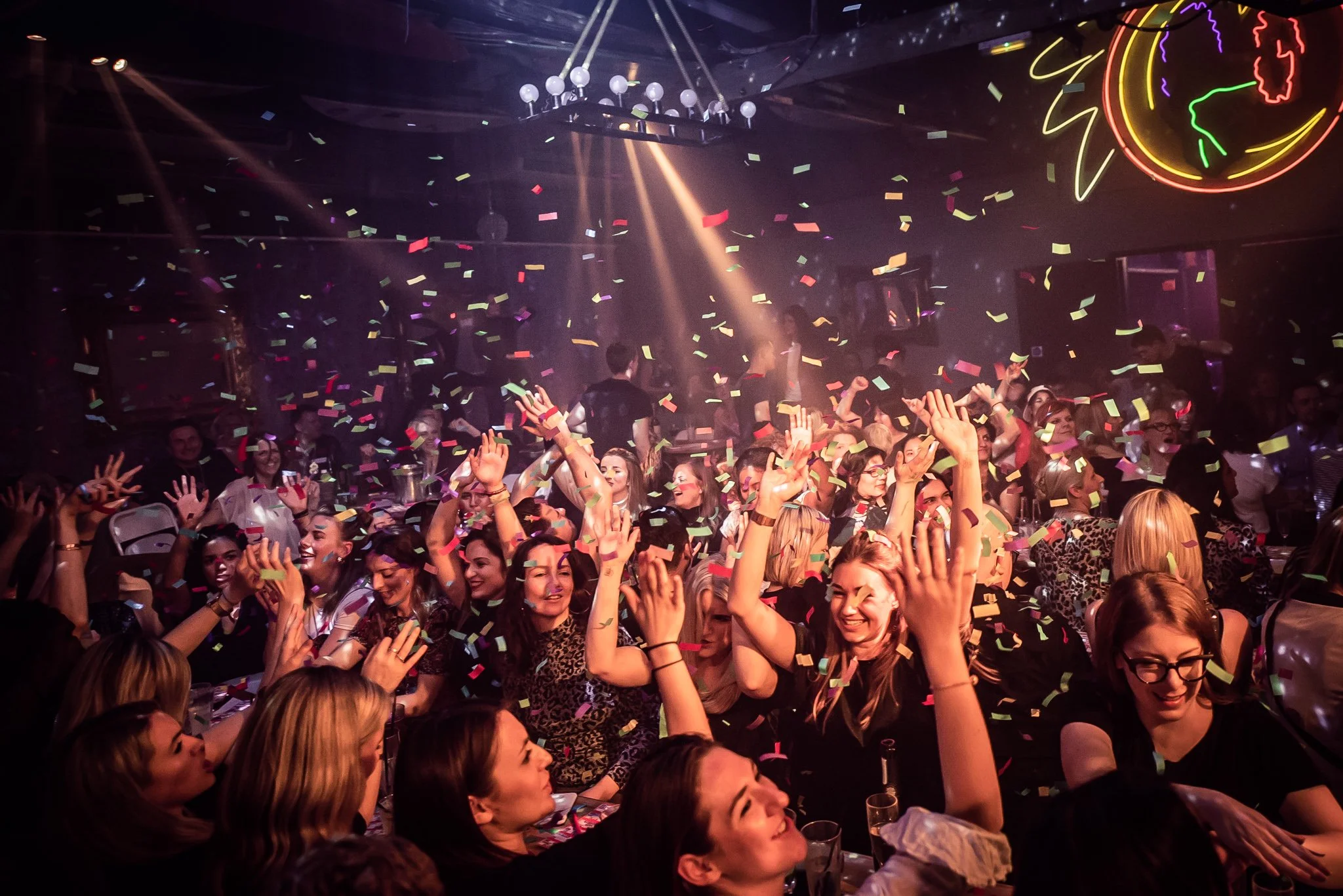 A crowded dance floor at a nightclub with many people celebrating, confetti falling, and colorful neon lights on the ceiling.