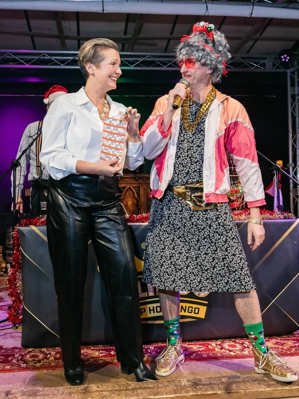A woman in a white shirt and black leather pants is smiling and holding a bingo card while talking to a man dressed in colorful, eccentric festive attire, including a curly gray wig, glasses, a gold chain, a patterned dress, and bright socks, on a decorated stage.