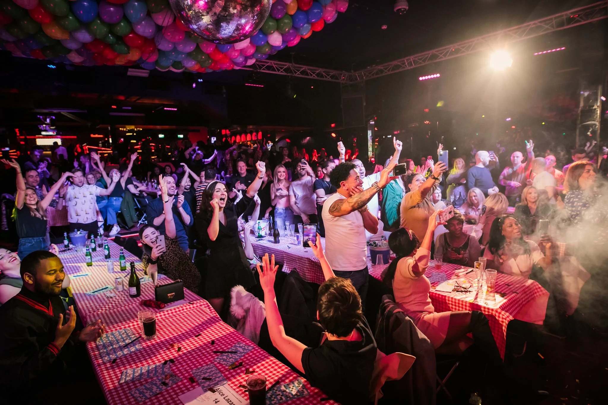 A large crowd of people at a lively party or concert, dancing and celebrating under colorful balloons and a disco ball, with bright stage lighting and a vibrant atmosphere.