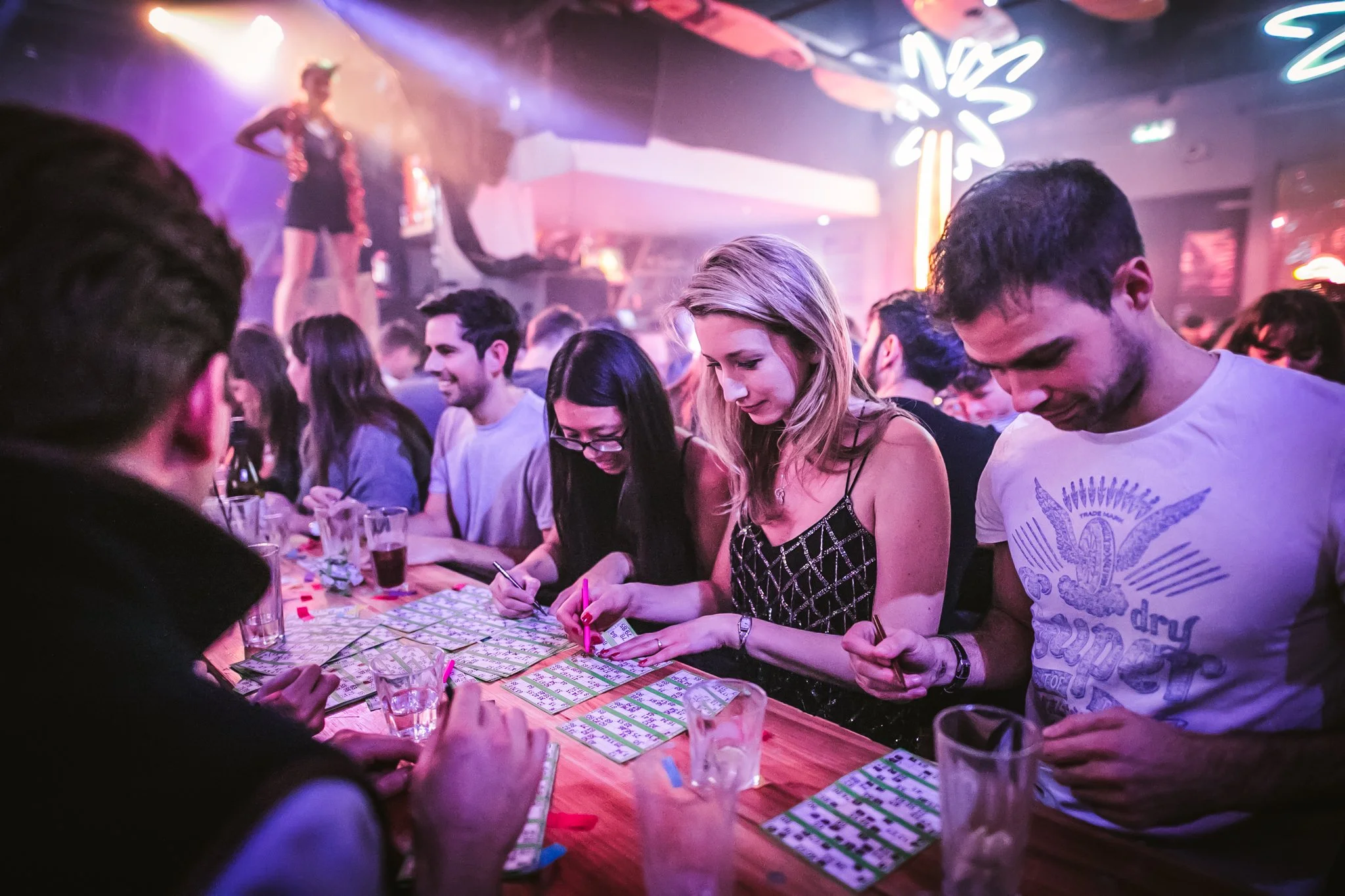 People gathered around a table playing bingo at Blingo, with a performer on stage and neon signs in the background.