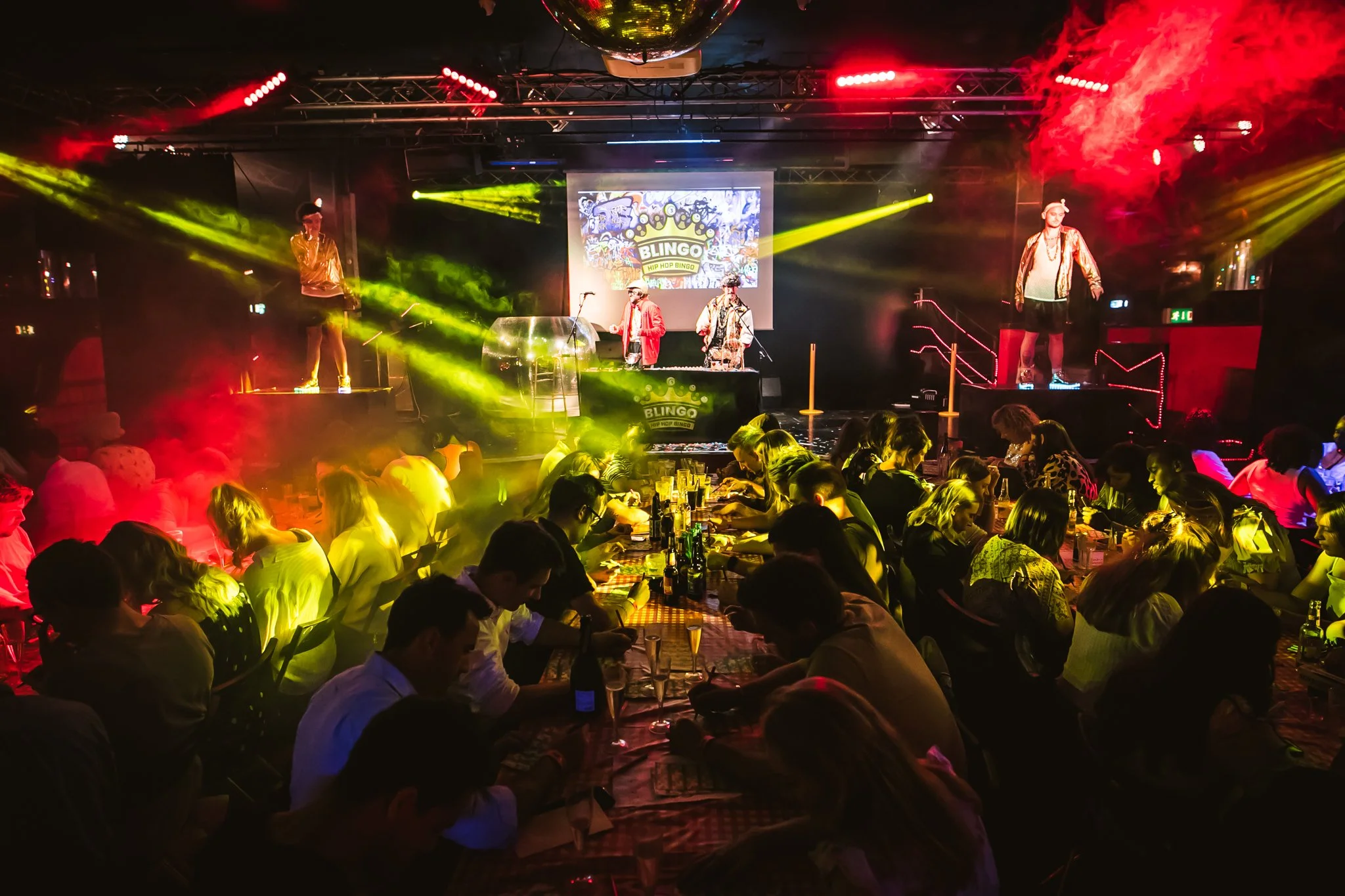 People sitting at tables at Blingo Bingo in a dimly lit venue with colorful stage lights, a DJ set, and a large screen displaying a 'Bingo' sign. The scene suggests a lively party or event with a dance floor.
