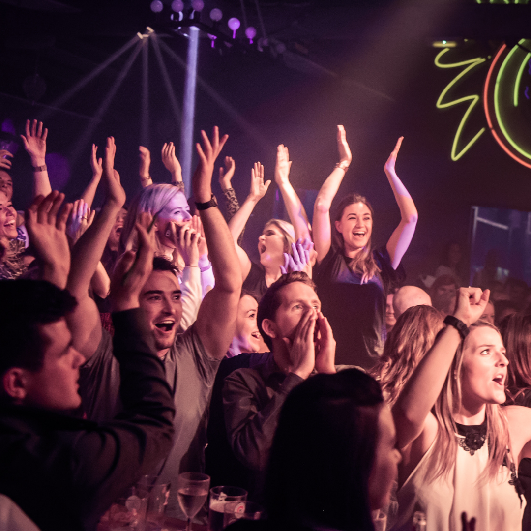 Group of people at a concert or party, smiling, cheering, with hands raised, illuminated by stage lights.