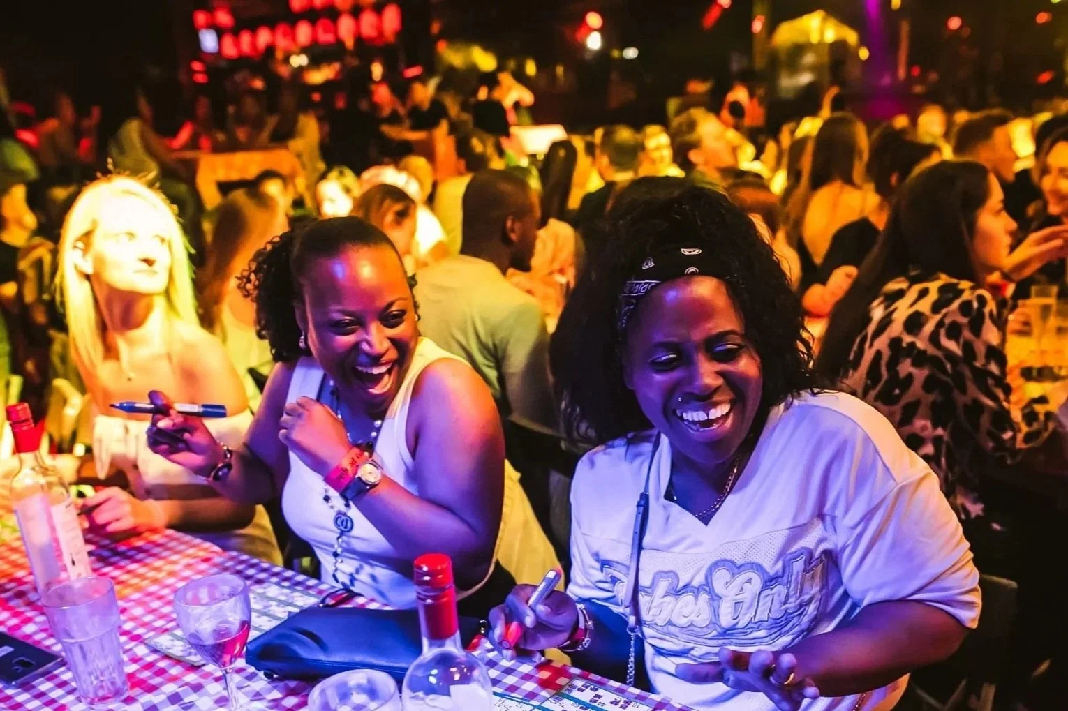 Two women laughing and enjoying drinks at Hip Hop Bingo party, surrounded by a crowd of people in a dimly lit setting with colorful lights.