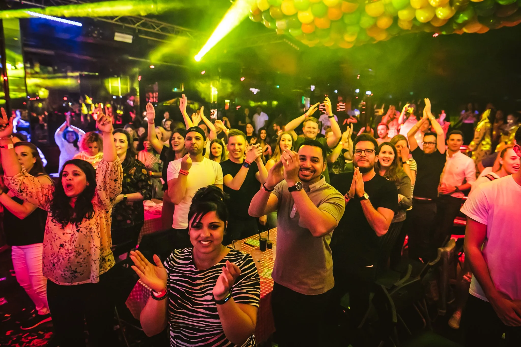 A large crowd of people enjoying a concert or party under colorful lighting and yellow balloons hanging from the ceiling.