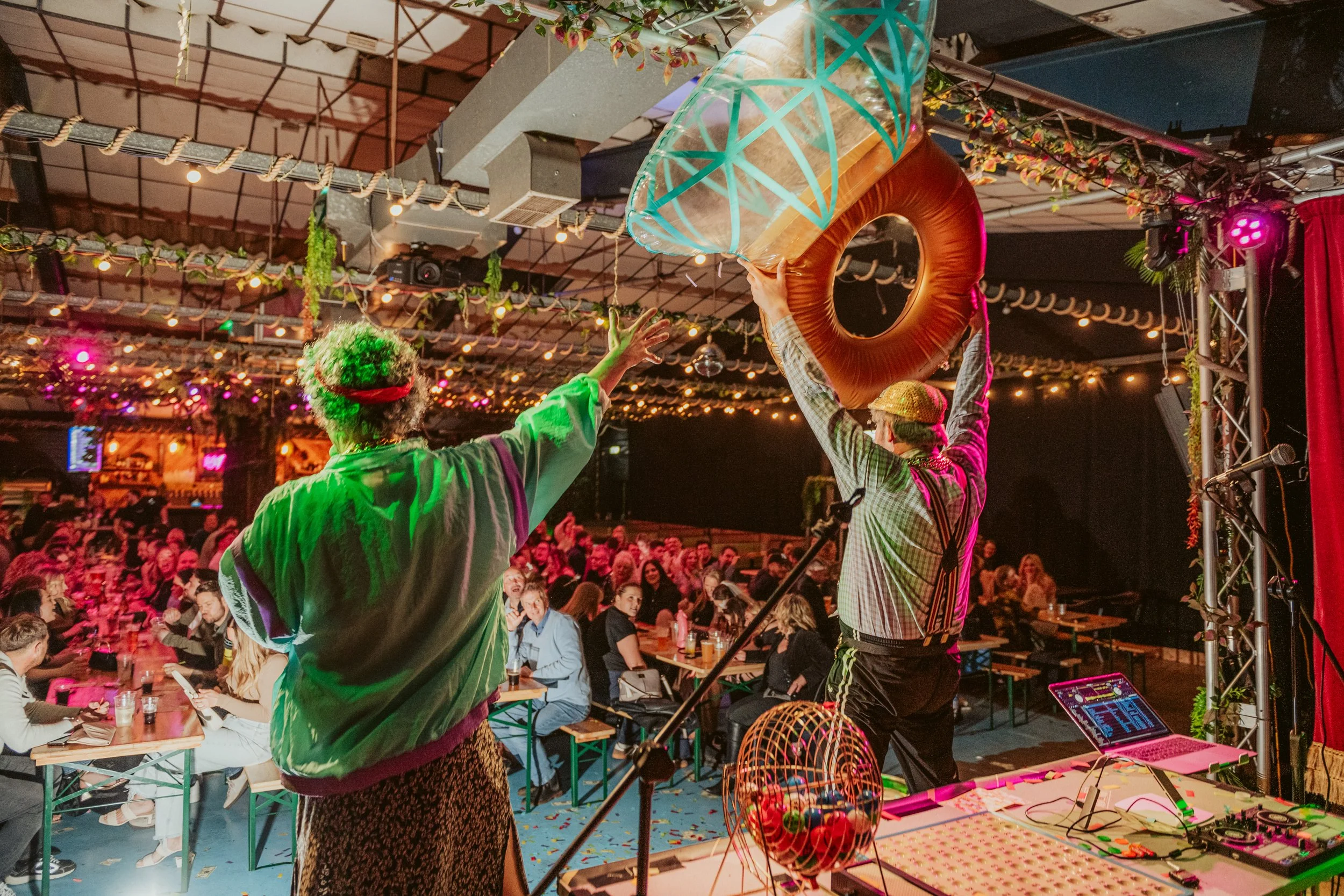 Two performers on stage at bingo with one holding a giant inflatable ring, is an indoor event with a crowded audience, colourful lighting, and decorative string lights overhead.