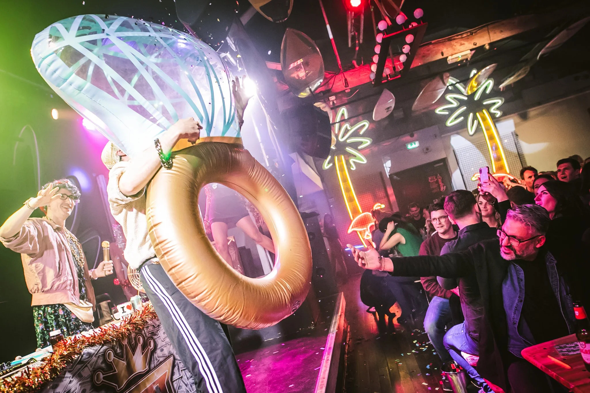 People in a nightclub dancing and taking pictures, with a colorful neon sign and palm tree decorations in the background.