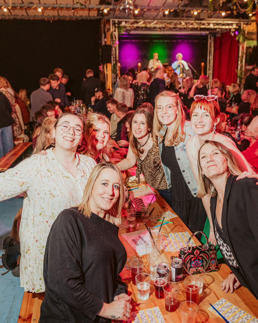 Group of six women smiling and posing for a photo at a party or event, with a stage and performers playing music in the background, drinks on the table, and a lively, festive atmosphere.