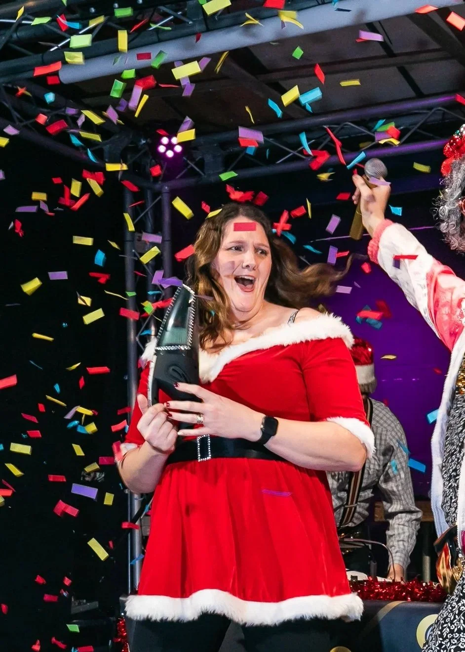 A woman dressed in a Santa Claus-inspired red dress with white fur trim is celebrating with colorful confetti falling around her, holding a microphone, and appearing excited and joyful.