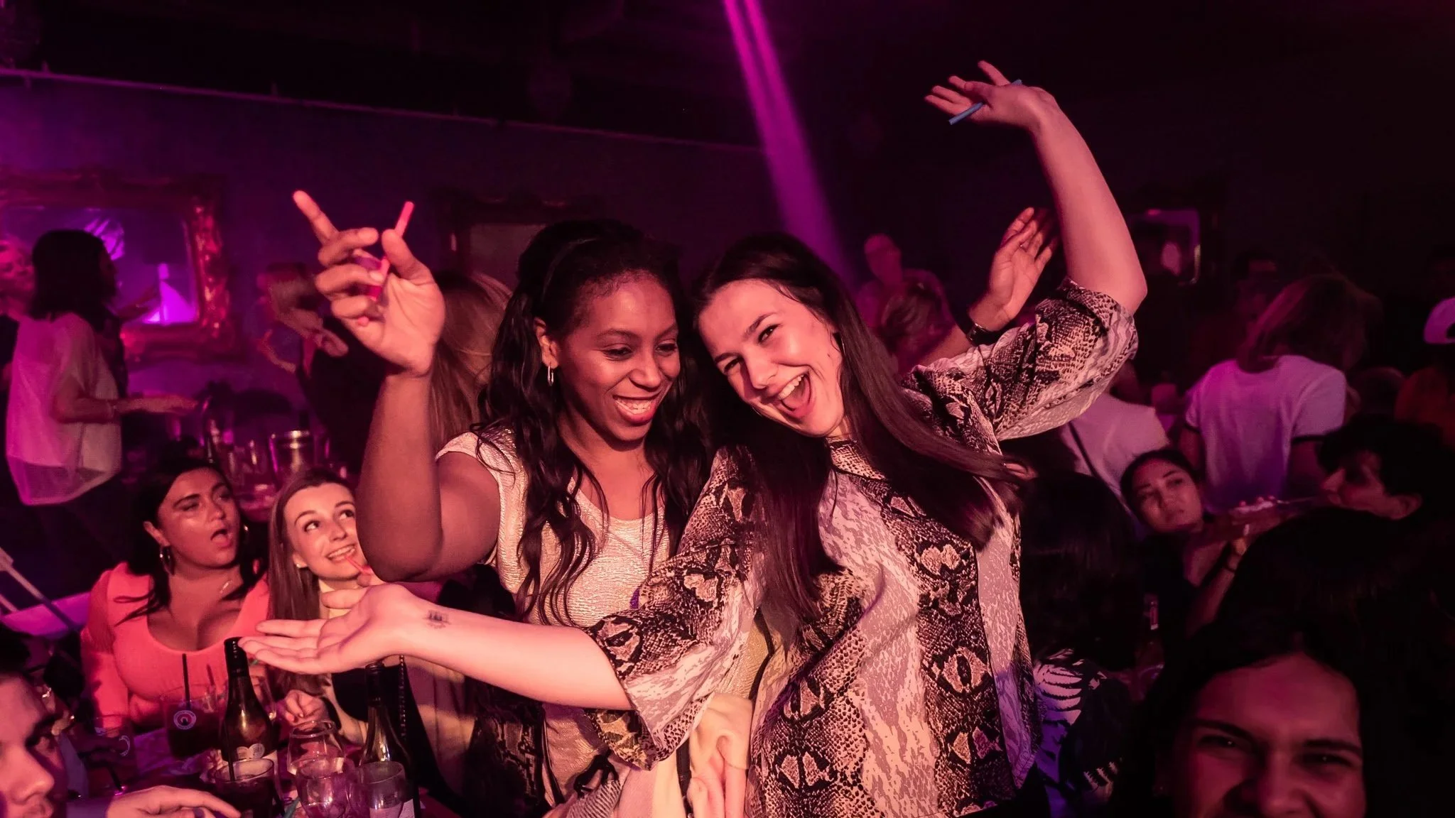Two women dancing and smiling at Hip Hop Bingo with purple lighting, surrounded by other people, drinks, and party atmosphere.