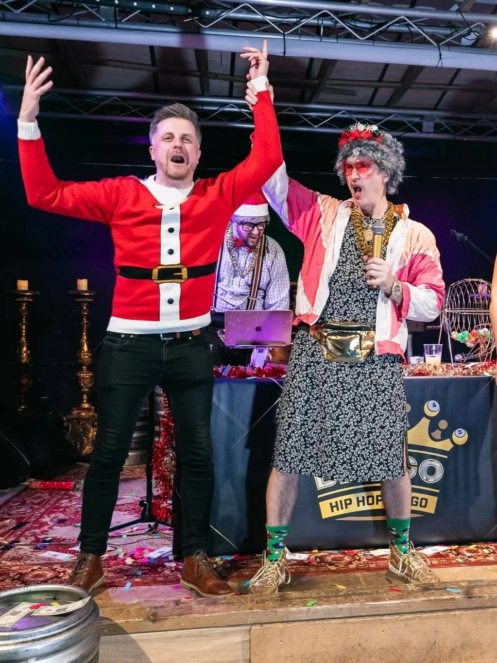 Two men dressed in humorous Christmas costumes on stage, one as Santa Claus and the other as an eccentric character wearing a floral dress, holding a microphone, with a DJ in the background. The scene appears festive with confetti and decorations.