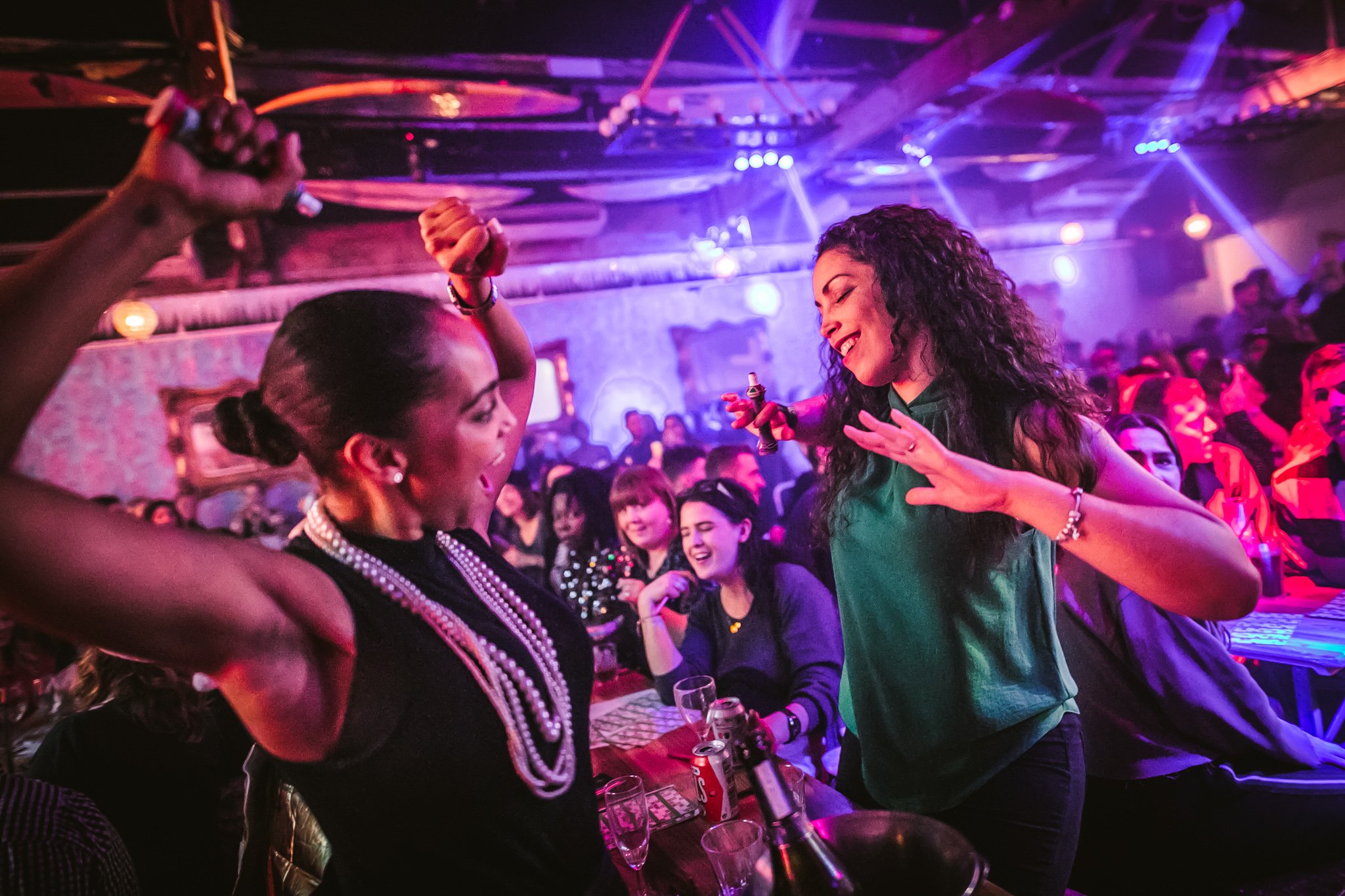 A lively scene in a nightclub with women dancing and socializing. The woman on the left is wearing a black dress and pearl jewelry, and is holding a bottle. The woman on the right with curly hair is wearing a green top and is smiling while dancing. The background shows other patrons, purple and pink lighting, and a busy party atmosphere.