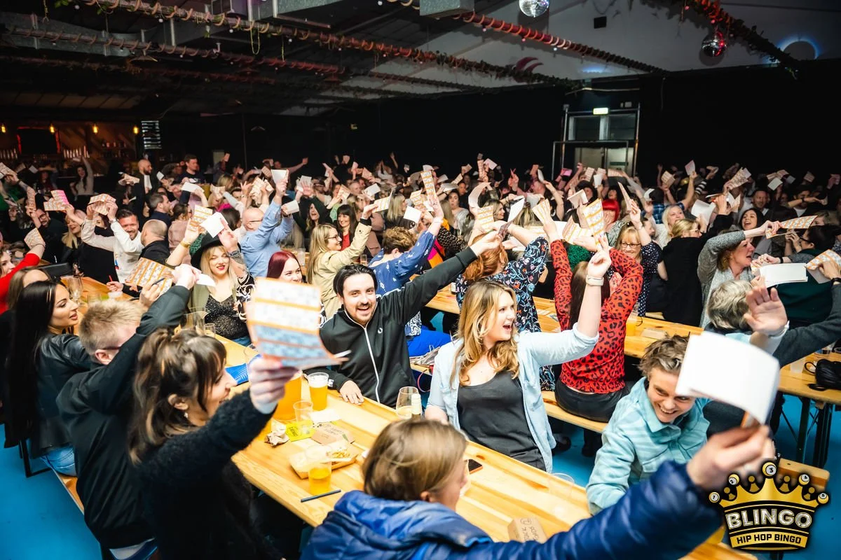 Large group of people at a party or event, holding up bingo cards and celebrating, with some smiling and cheering.