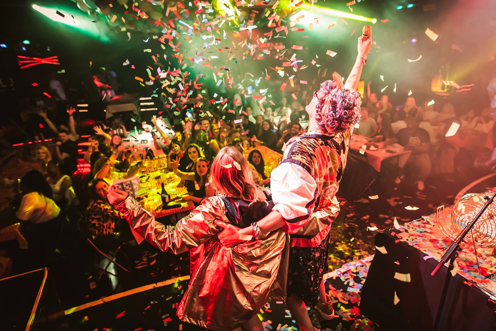 Person on stage holding microphone high amidst colorful confetti and vibrant stage lights, audience cheering in a lively indoor party setting.