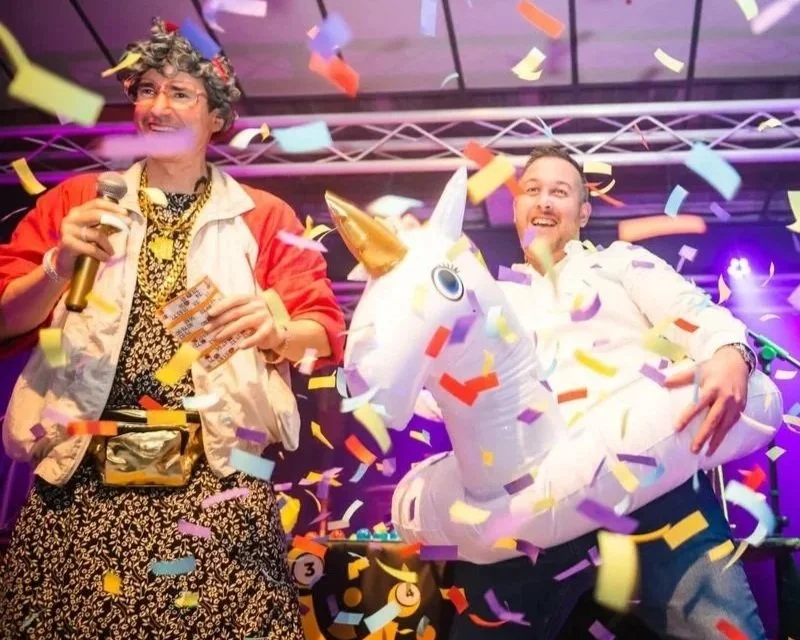 Two men celebrate winning bingo on stage with confetti, one dressed in a leopard print outfit with a gold chain and holding a microphone, the other in a white shirt riding a large inflatable unicorn.