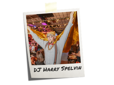 Dj Harry Spelvin of Hip Hop Bingo with glasses and a beard wearing a checkered shirt, raising his arms in celebration at an indoor event with colorful lighting and a crowd in the background.