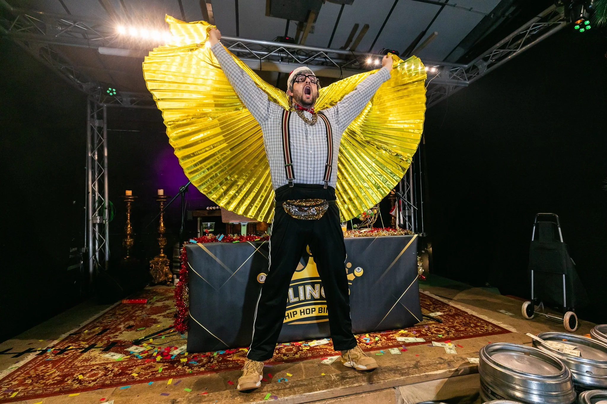A man on stage with a yellow fan-shaped prop, wearing glasses, plaid shirt, suspenders, and a necklace, shouting or singing with arms raised, surrounded by confetti and Christmas decorations.