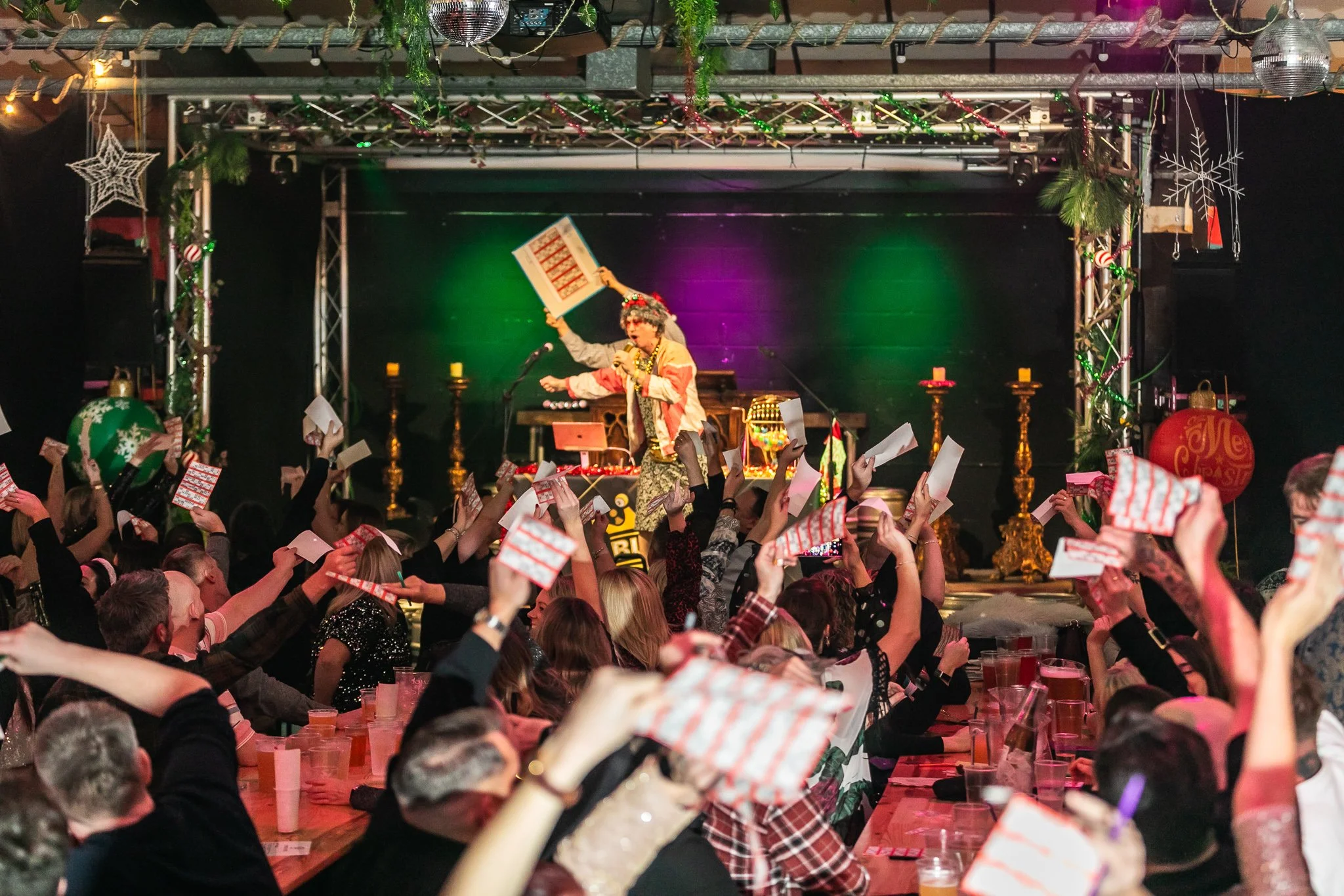 A performer dressed in festive attire is singing into a microphone on stage at a Christmas celebration, surrounded by decorated holiday ornaments and an enthusiastic audience holding up greeting cards or papers.