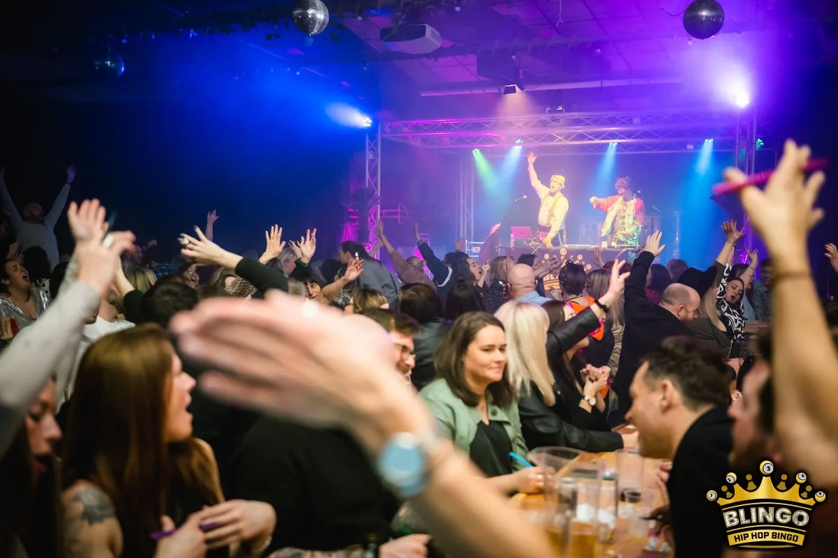 Crowd dancing and enjoying a London bingo night on stage, colourful lights, and a lively atmosphere.