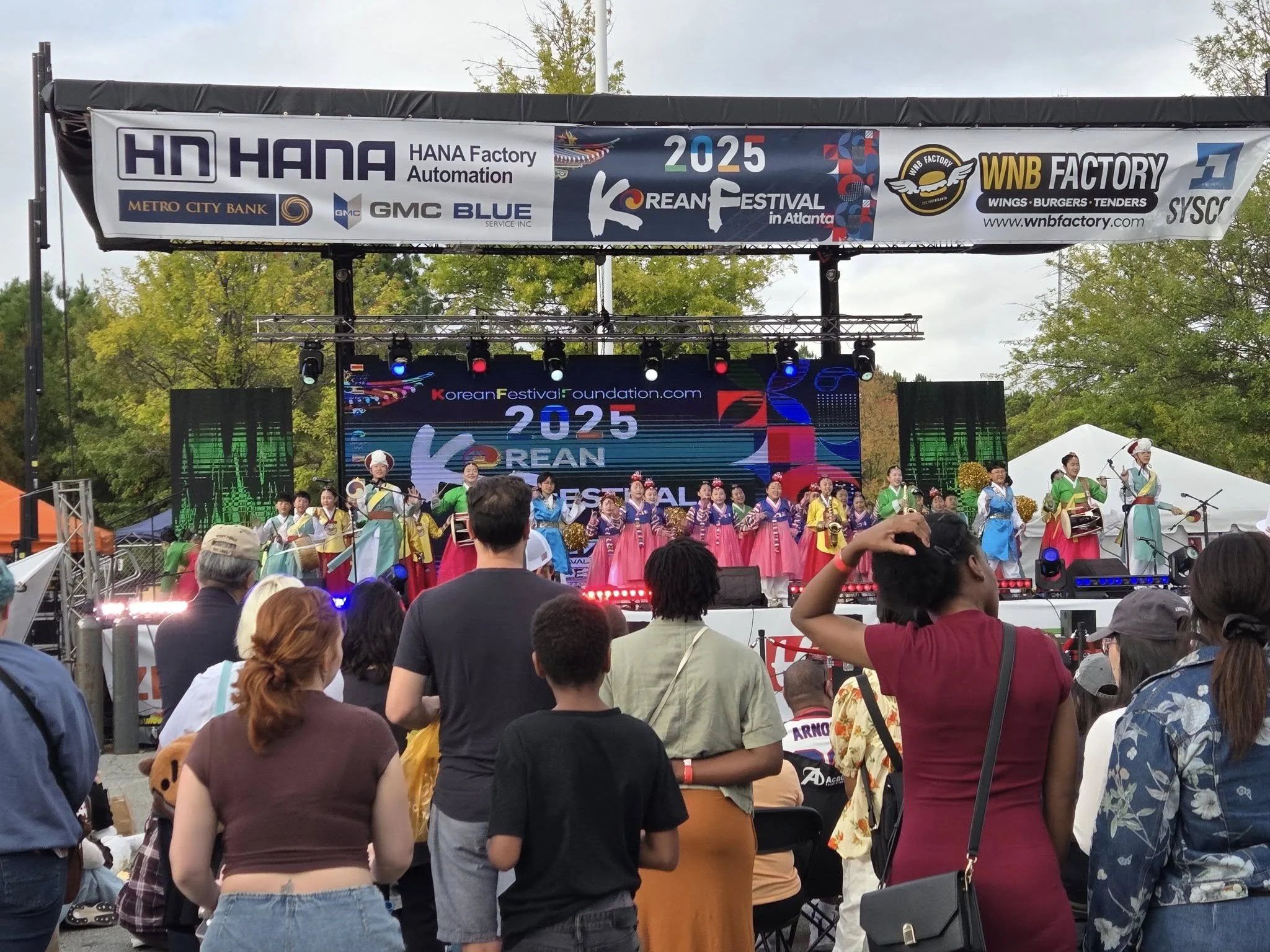 한인 축제에서 전통 의상을 입은 무용단이 무대에서 공연하는 모습입니다. 관객들이 공연을 감상하며 서서 보고 있습니다.