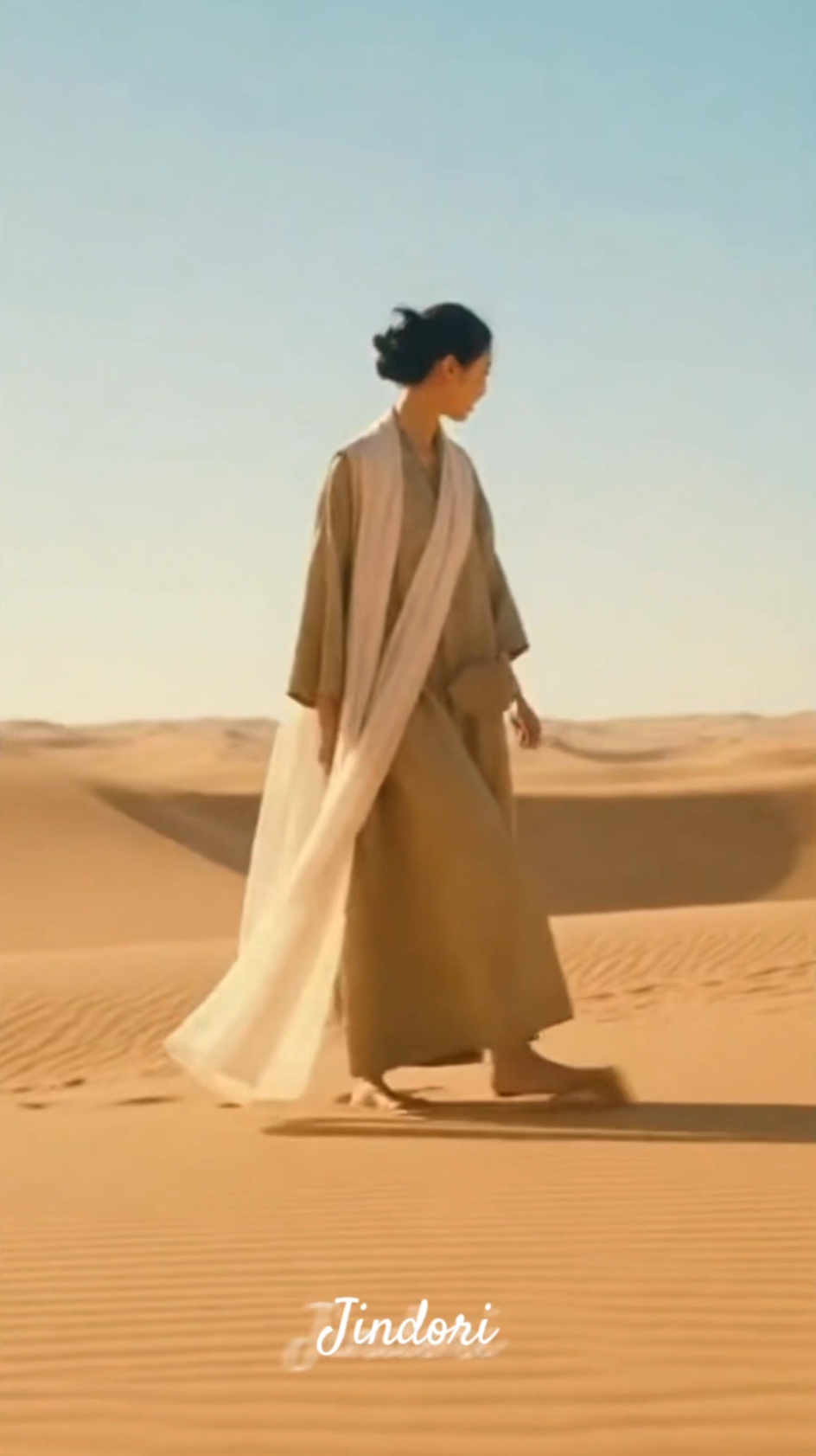 사막을 배경으로 한 여성의 모습, 갈색 옷을 입고 걷고 있음, 하늘은 맑음.