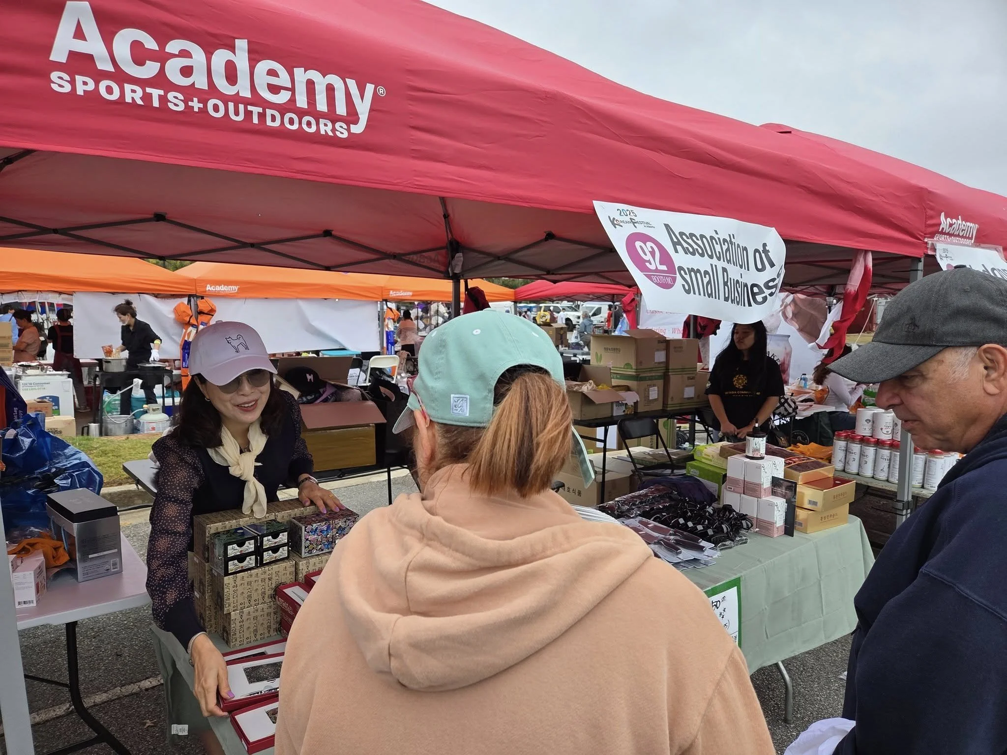 아카데미 스포츠 아웃도어 텐트 아래의 판매 부스에서 사람들이 제품을 구매하고 있으며, 배경에는 'Association of Small Business'라는 표시가 붙어 있다.