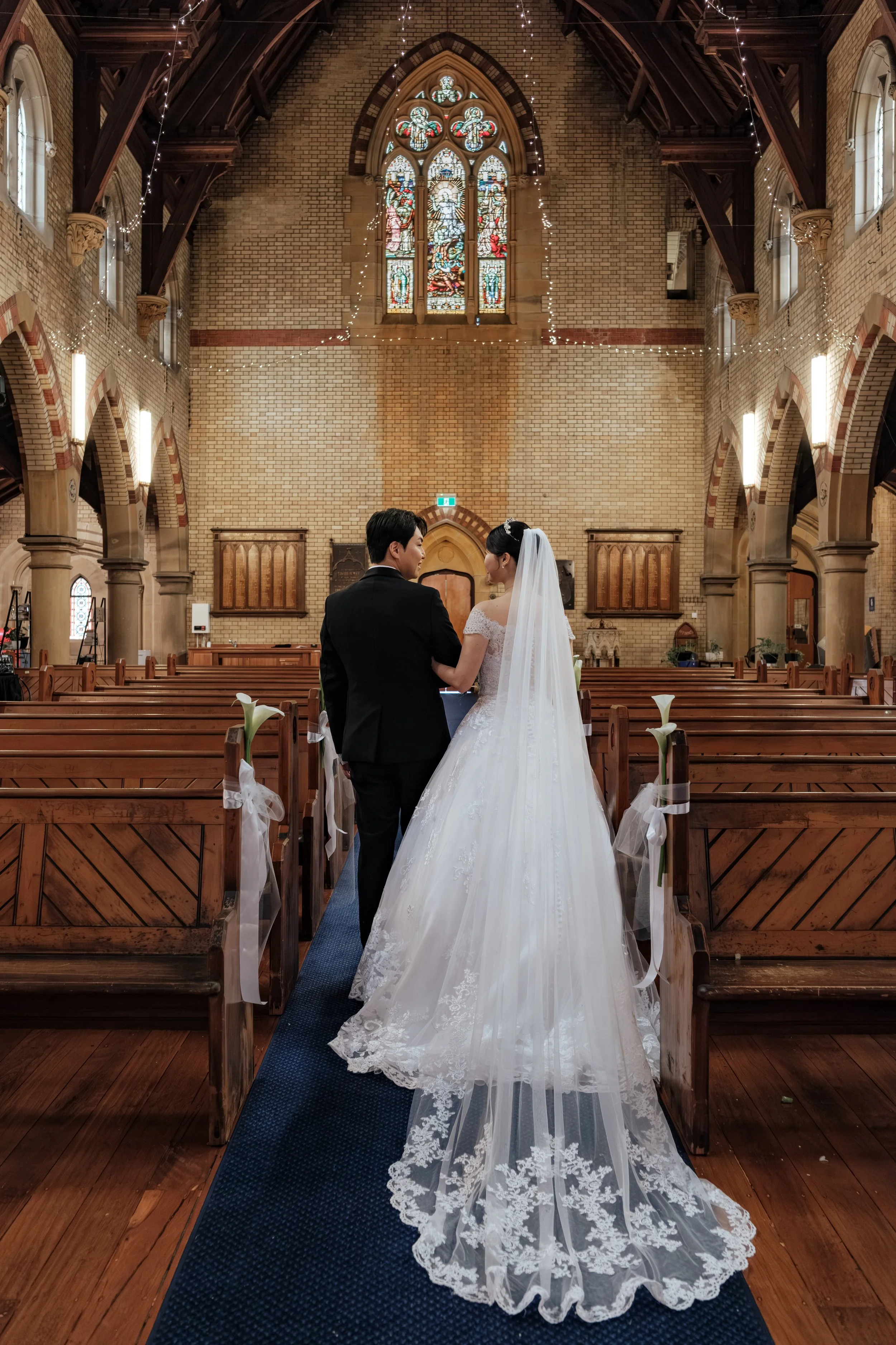 Bride and groom holding hands in church during wedding ceremony, with pews decorated with white flowers and ribbons, stained glass window above, and warm lighting.