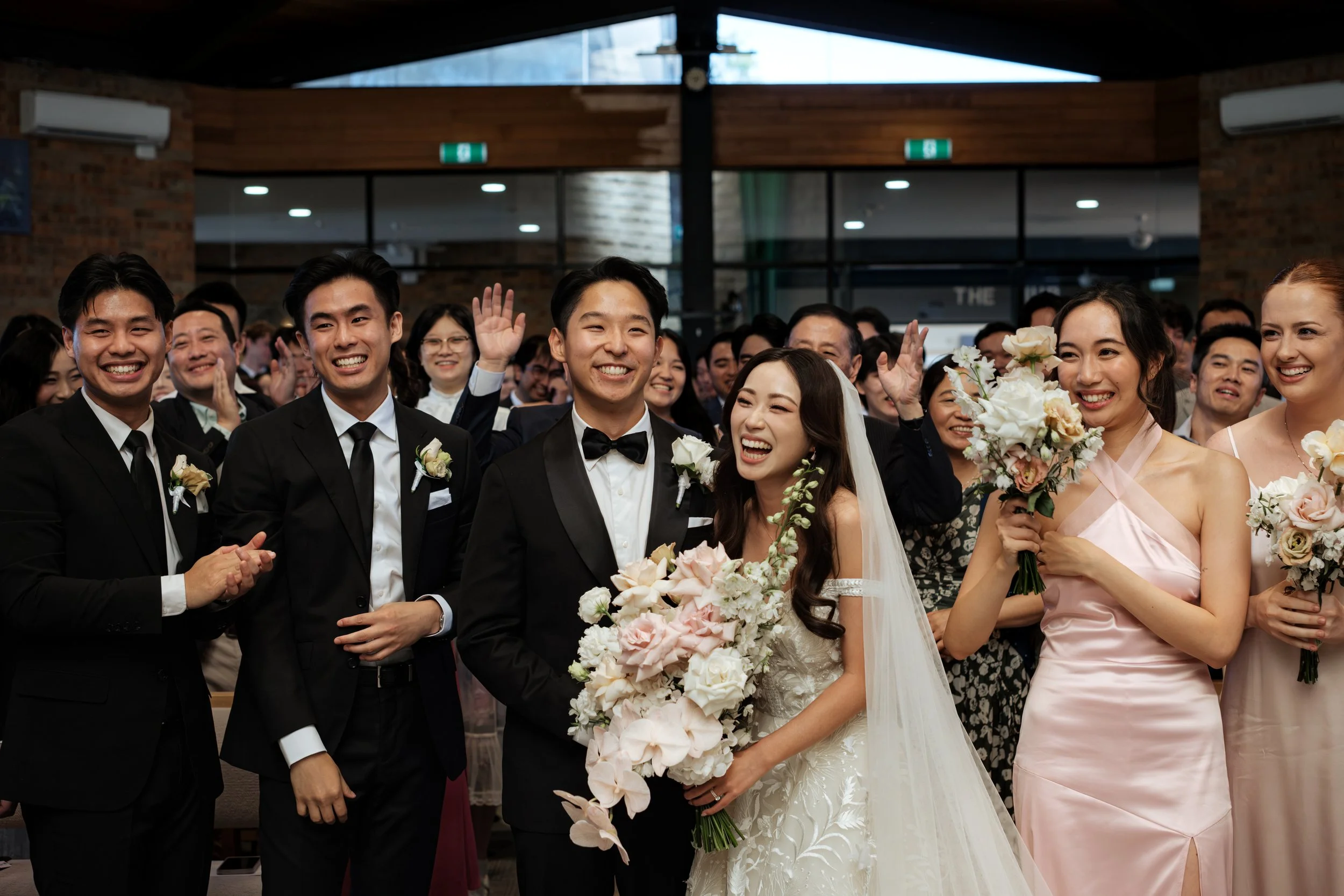 Wedding celebration with smiling bride and groom surrounded by wedding party and guests in a bright indoor venue.