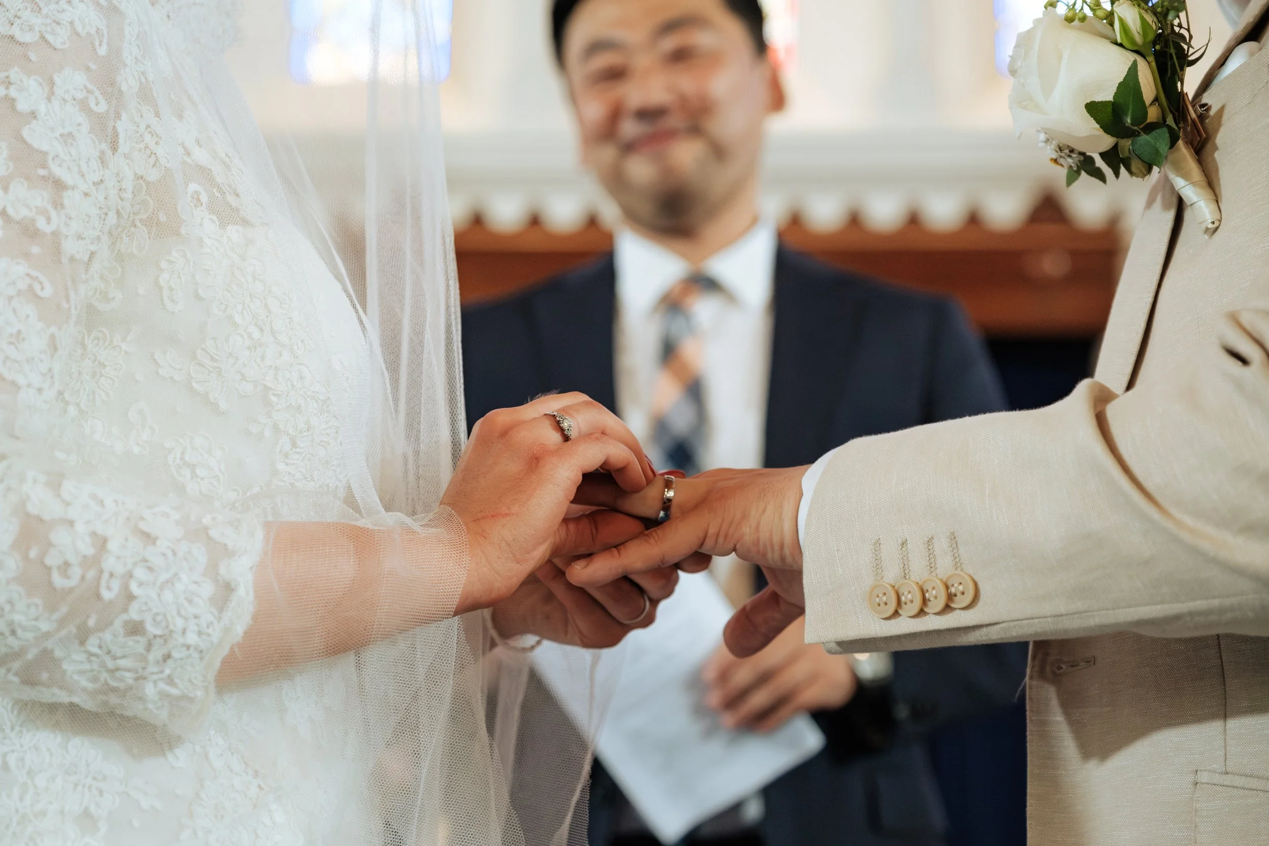 Close-up of a wedding ceremony where a couple is exchanging rings, with a officiant smiling in the background. The bride's hand, with a wedding ring, is being placed onto the groom's finger. The bride wears a lace dress with a veil, and the groom wea