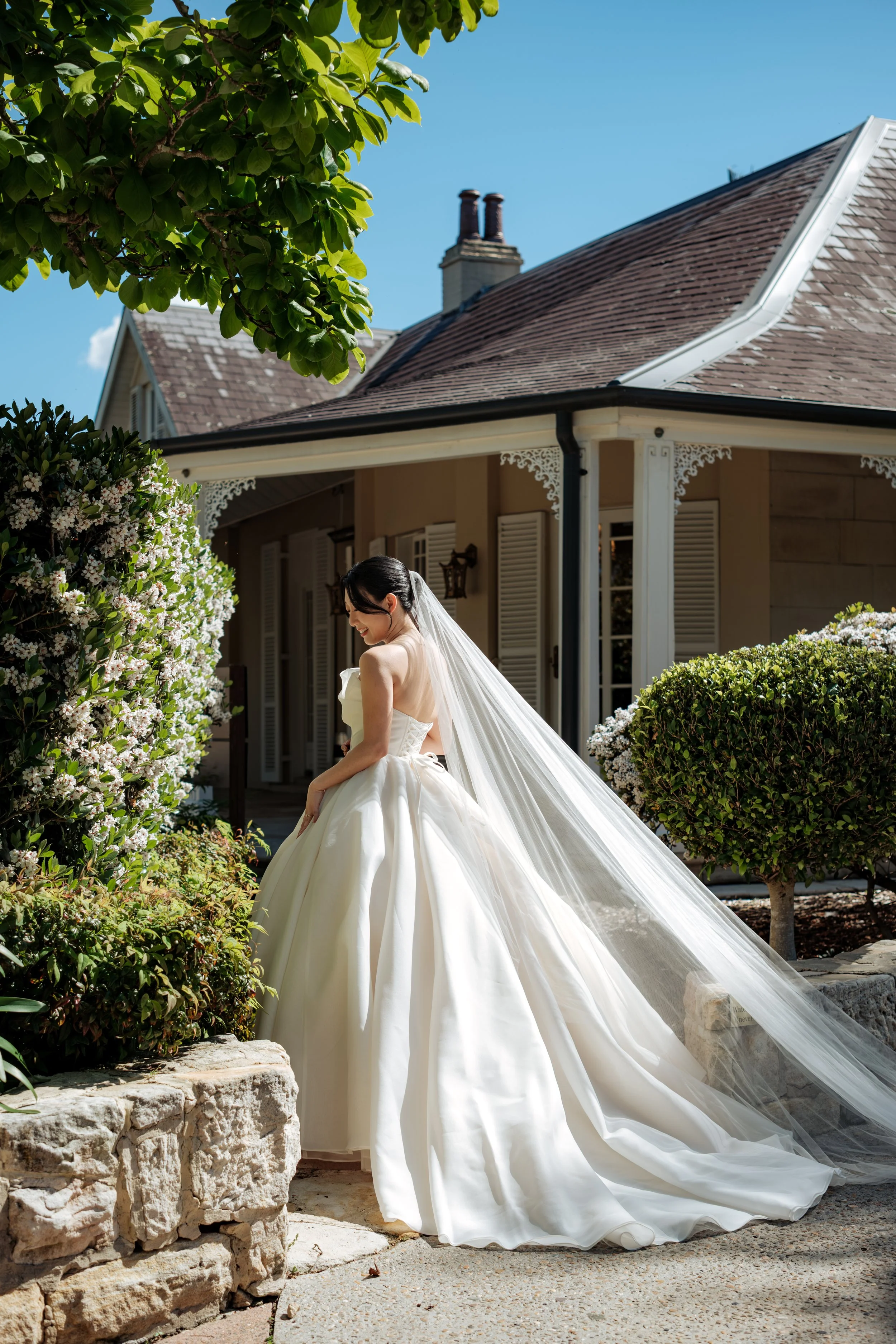 Bride in a white wedding gown with a long veil standing outside a house with white shutters and surrounded by flowering bushes and trees on a bright sunny day.