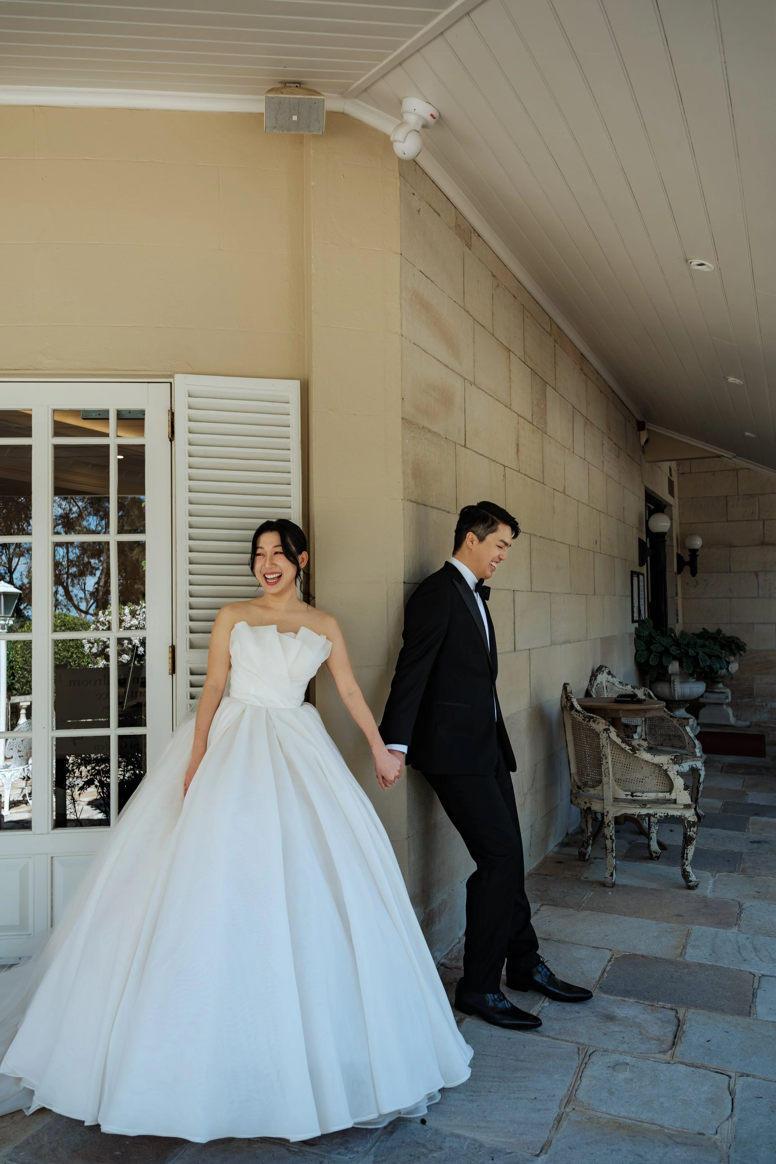 A newlywed couple in wedding attire standing against a beige stone wall, holding hands, smiling, outside a building with large glass doors and outdoor furniture.