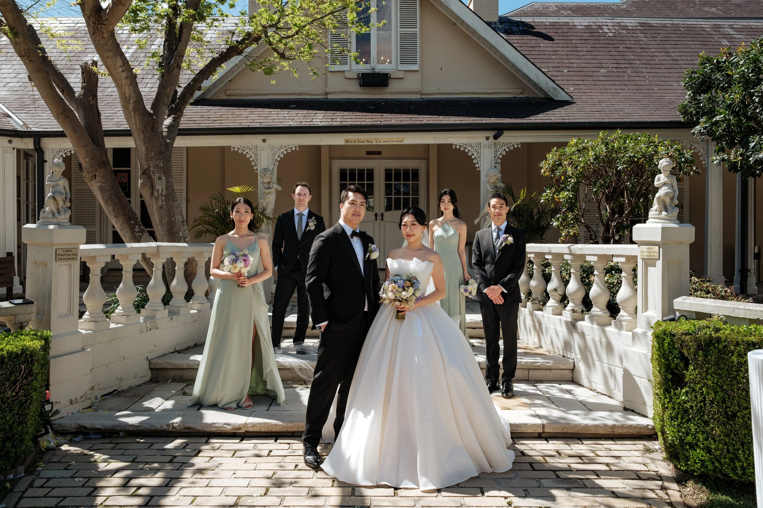 A bride and groom standing outside on steps with four wedding party members, two women and two men, in front of a house with charming Victorian details and statues.