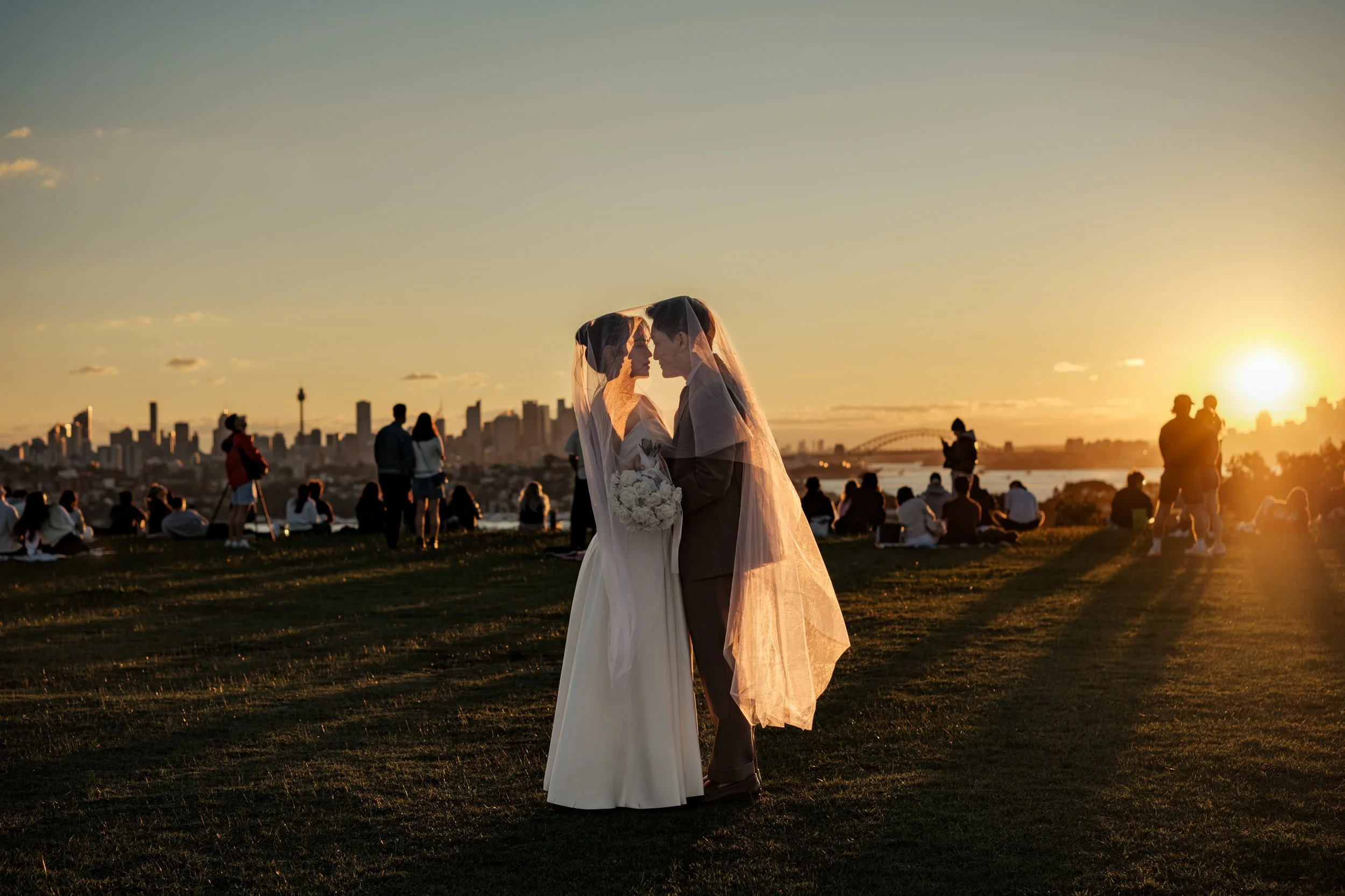 A bride and groom stand close, facing each other under a veil during sunset, with a city skyline in the background and people sitting and standing on a hill.